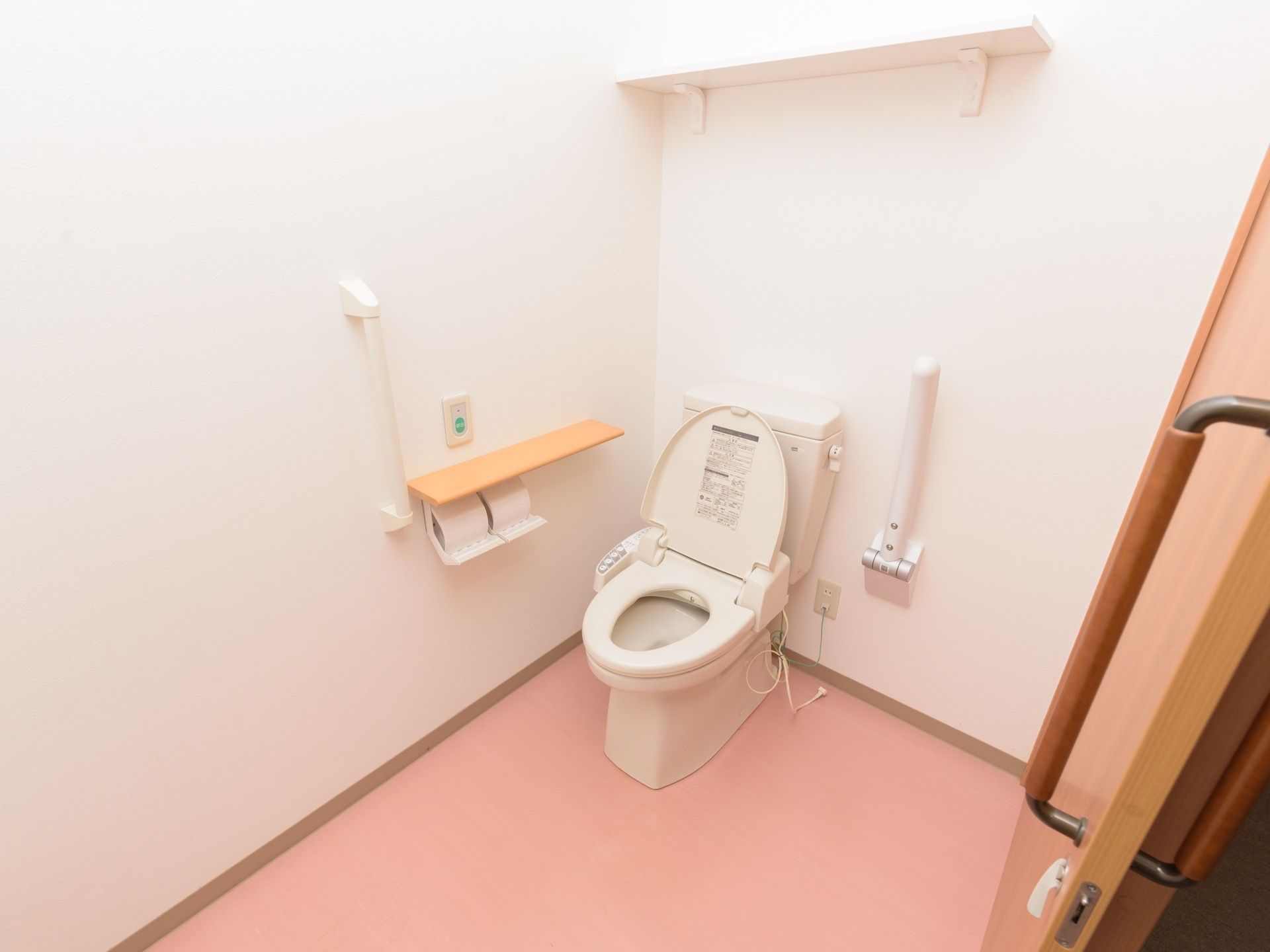 ピンク色の床の明るいトイレスペース。直線的なグリップや折りたたみ式のアームレストが便器周辺に設置されている。