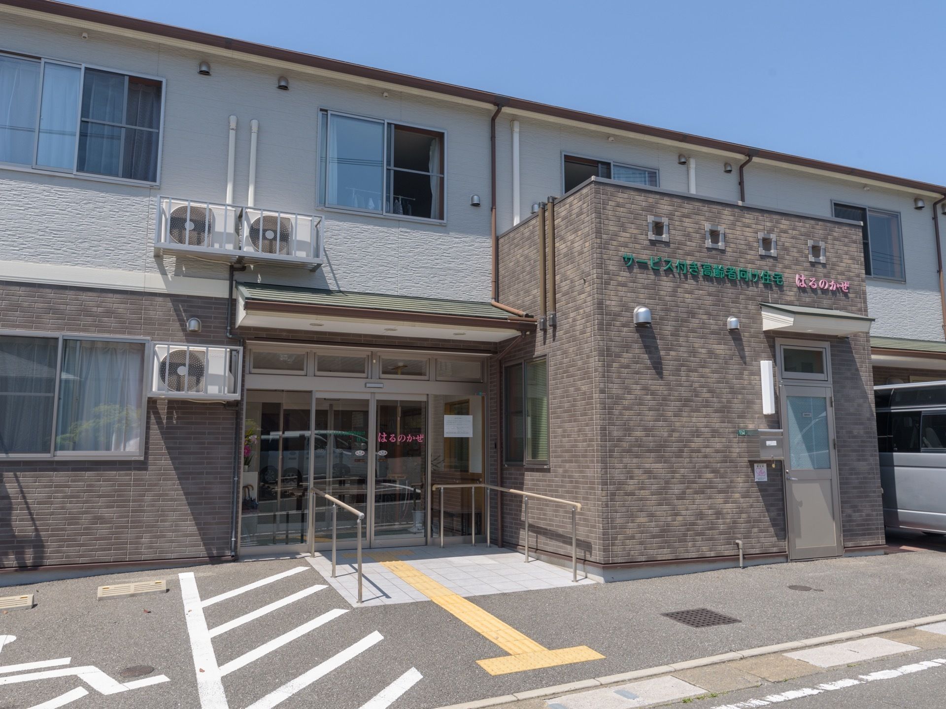 さっぱりとした外壁が施された２階建ての施設。建物の前はアスファルトで舗装されており、左側に優先駐車場が設けられている。