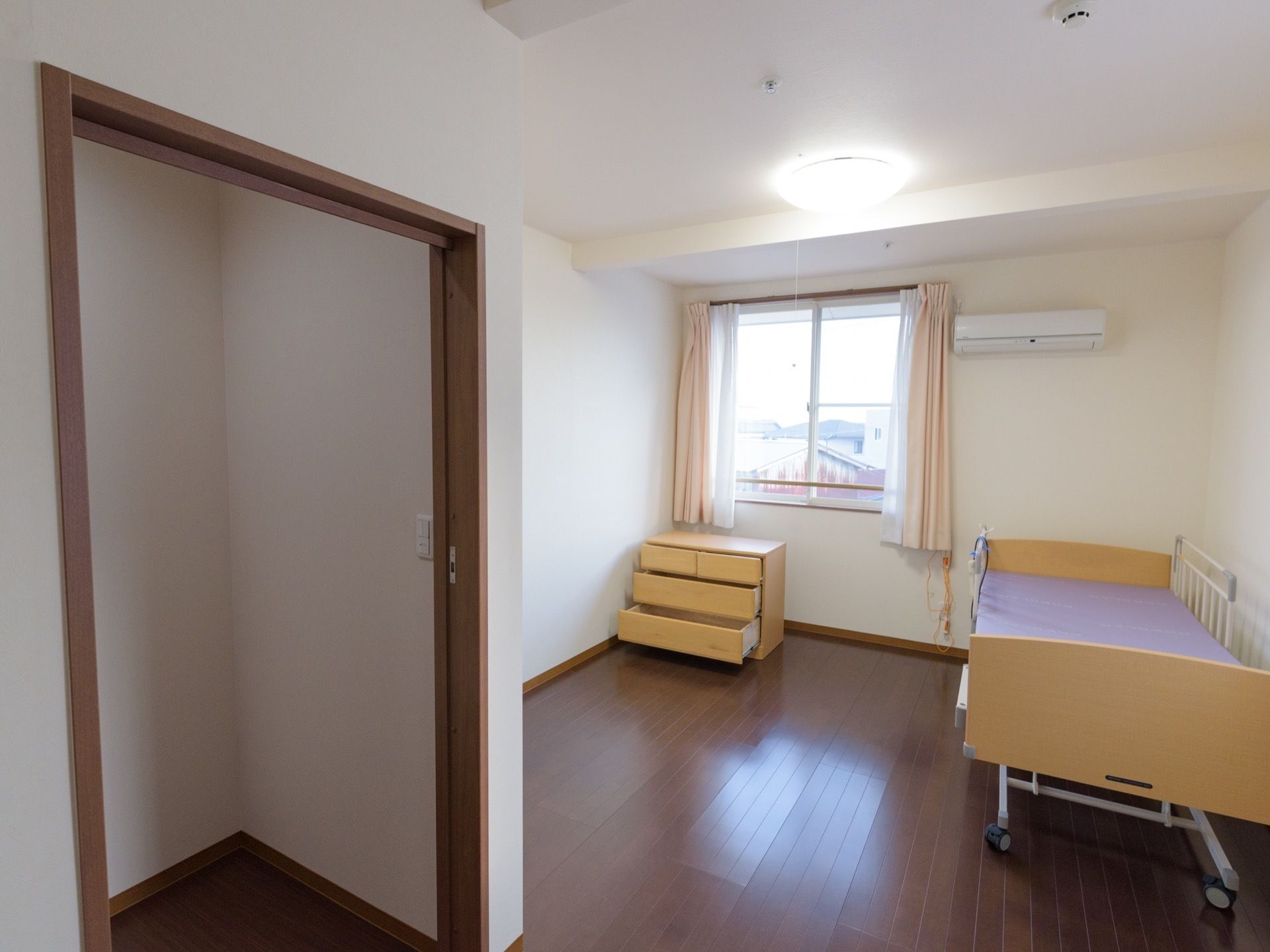 居室はフローリングで介護用ベッドと3段のタンスが置いてある。クーラーも設置してある。入口付近に収納庫もある。