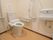 洗面台が備わった広々としたトイレである。可動式やＬ型手すり、呼び出しボタンが壁に設置されている。温水洗浄便座が便器に取り付けられている。