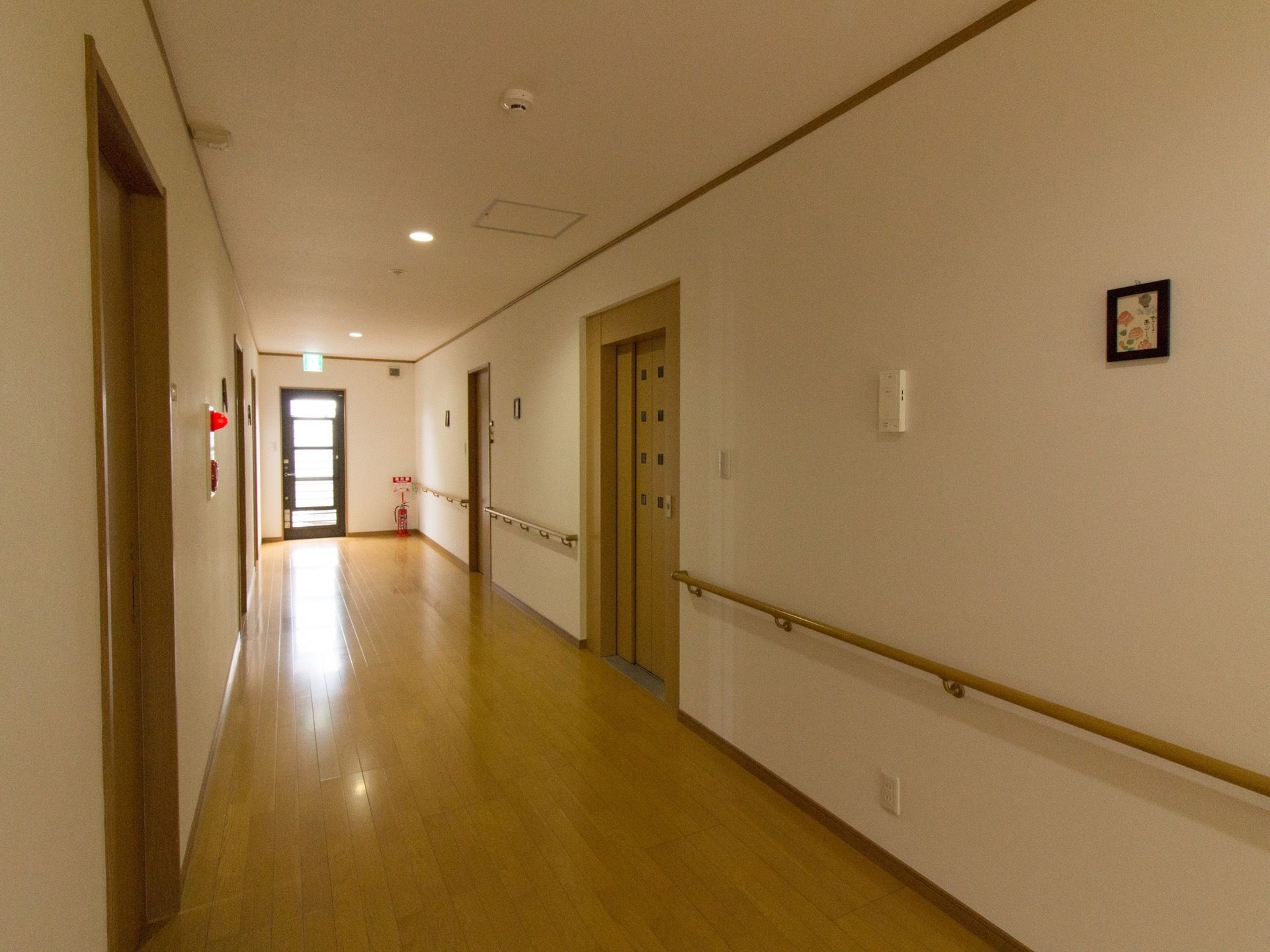 広々とした廊下の両側に居室やエレベーターが並ぶ。片側の壁には、手すりとコンセント、消火器が設置されている。