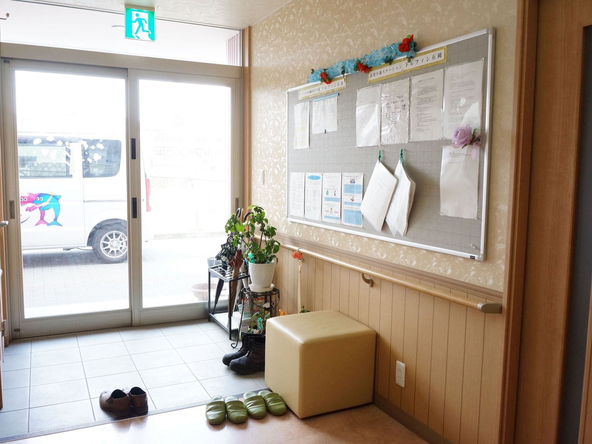 ガラスのスライドドアの入り口である。壁には大きな掲示板があり、書類が貼られている。スツールや傘立てが置かれている。
