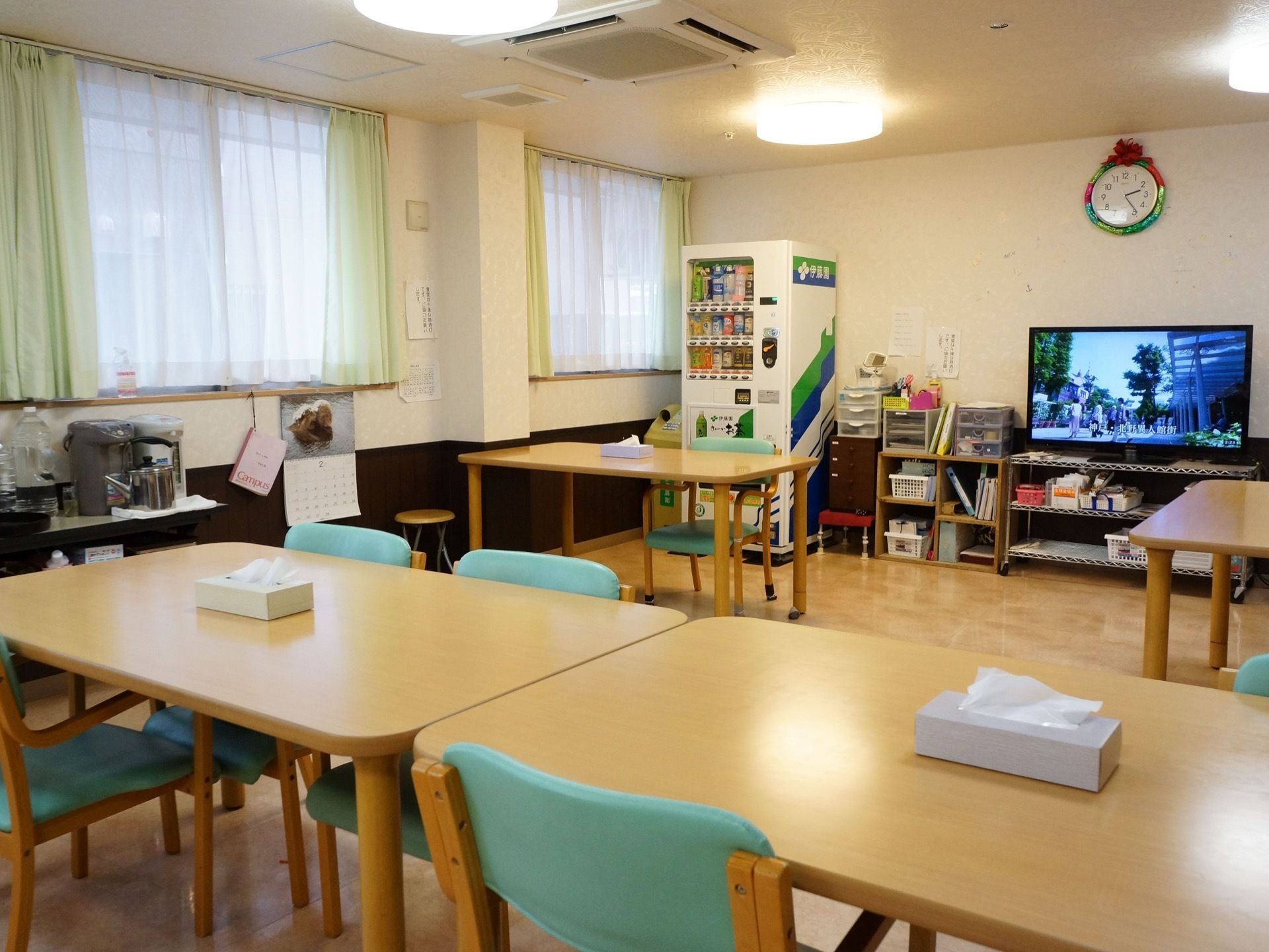 ダイニングテーブルや椅子が置かれている。壁際にはテレビや自動販売機が設置されている。窓の近くには、電気ポットが置かれている。