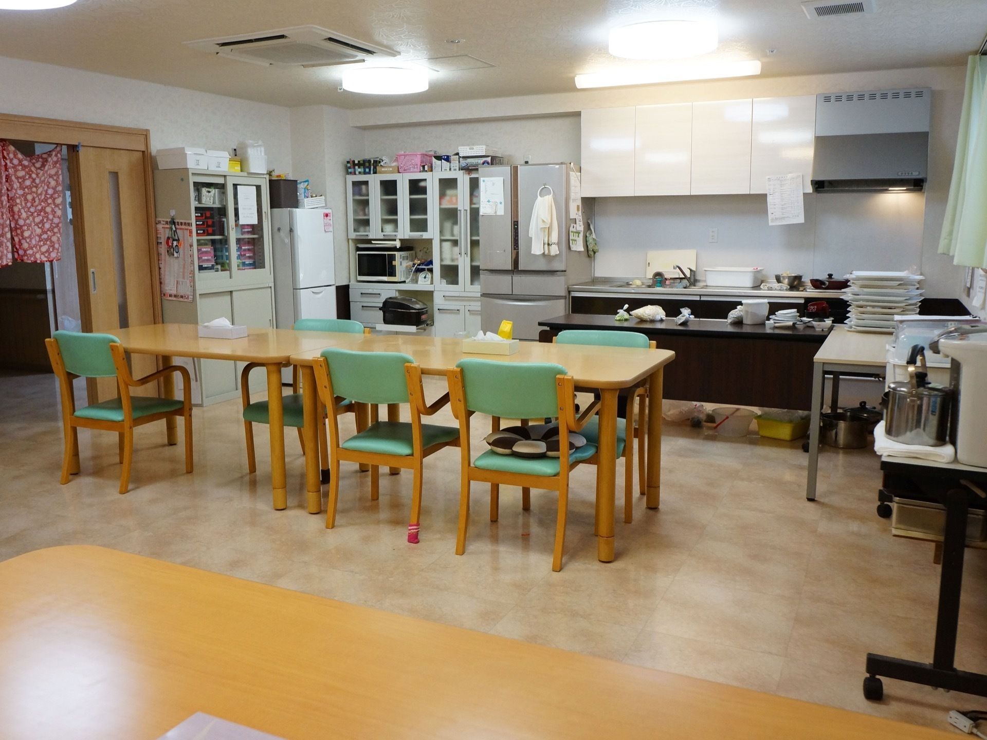 オープンキッチンが備わった食堂である。壁際には、冷蔵庫や食器棚が設置されている。椅子がセットされたダイニングテーブルが置かれている。