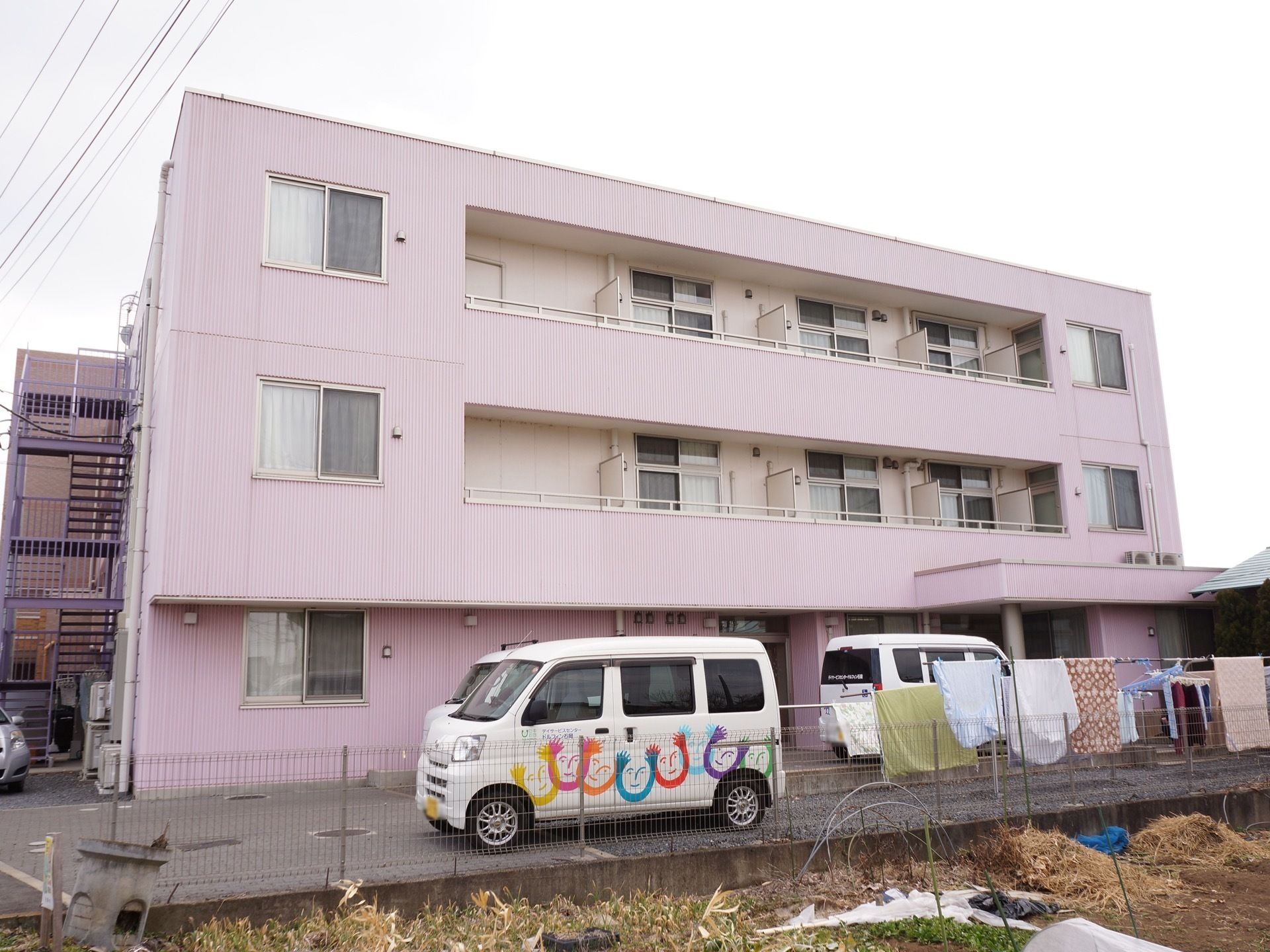 施設は、パステルピンクの２階建ての建物である。ベランダや外階段が設置されている。建物前に送迎車が停まっている。