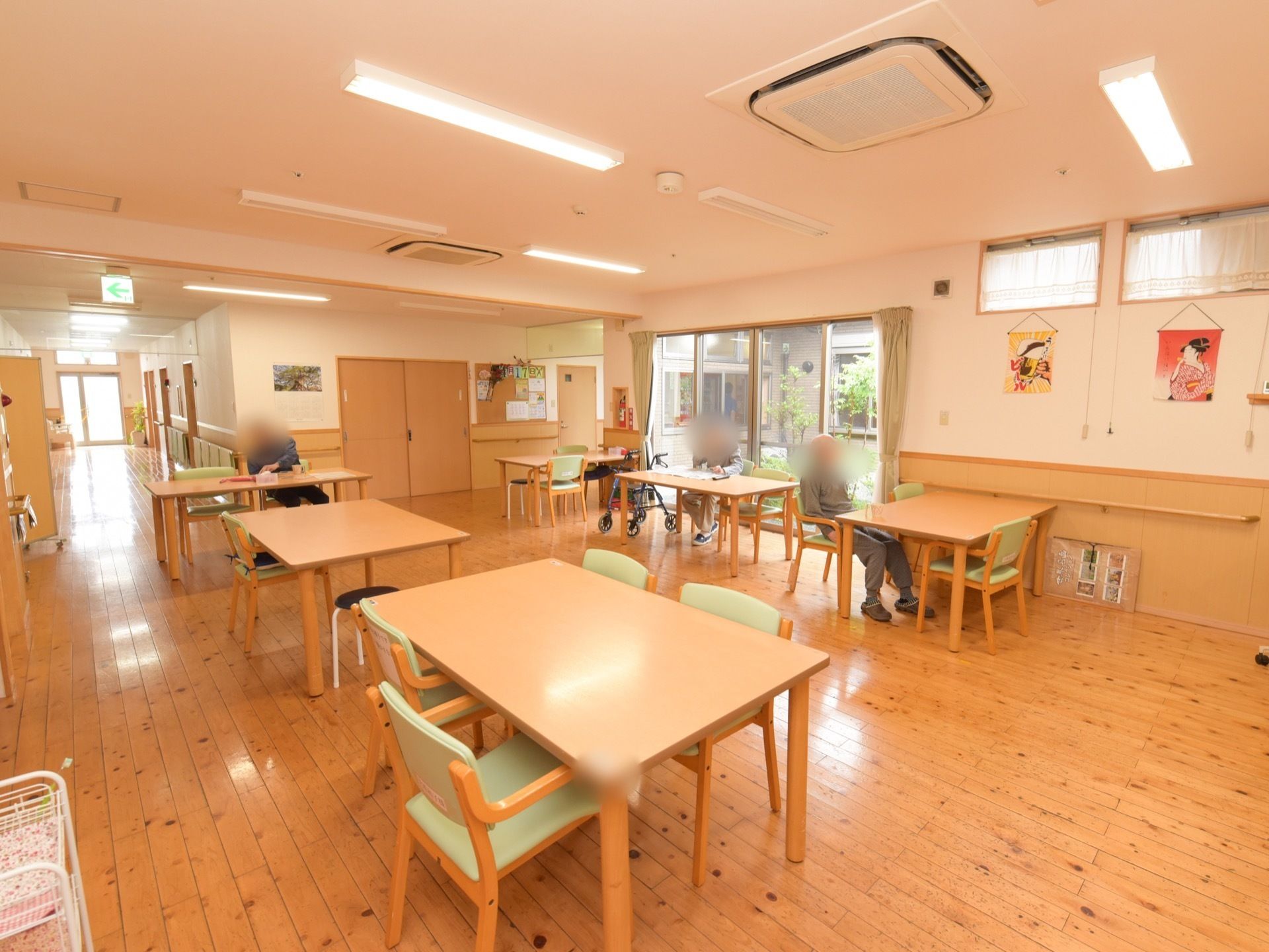 リビングは木製のフローリングで、数名の入居者様が椅子に座っている。壁には浮世絵のタペストリーが飾られている。