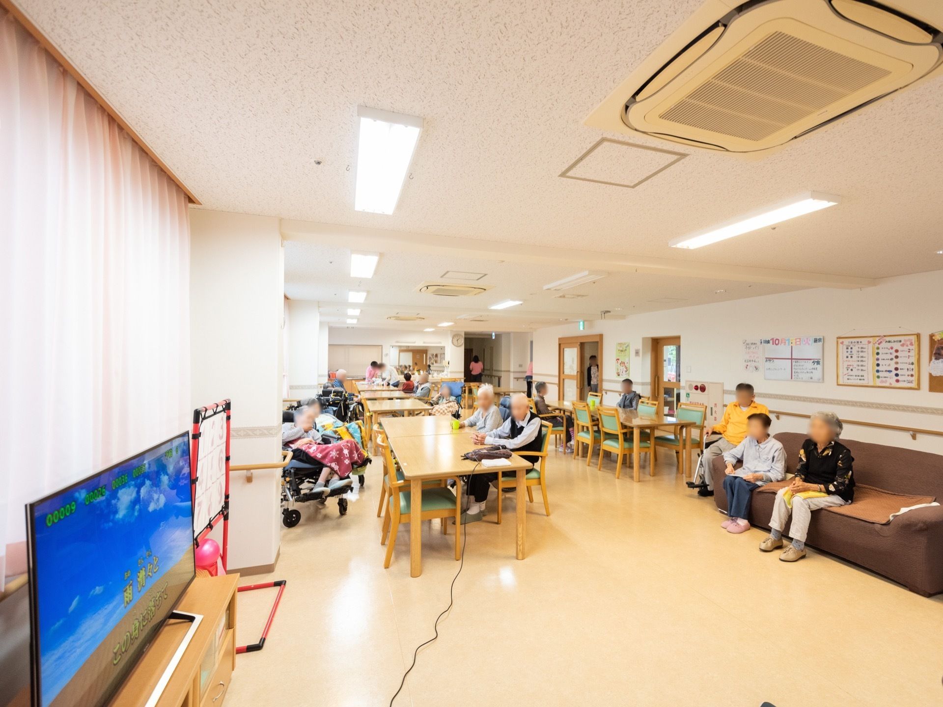 食堂に多くの入居者様が集まっていて、ソファや椅子、車椅子に座っている。窓際にあるテレビがついている。
