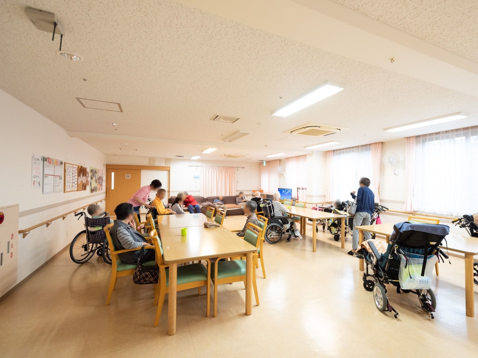 大きな窓が並んでいる明るい食堂がある。入居者様が大勢集まってテーブルについている。スタッフがお世話をしている。