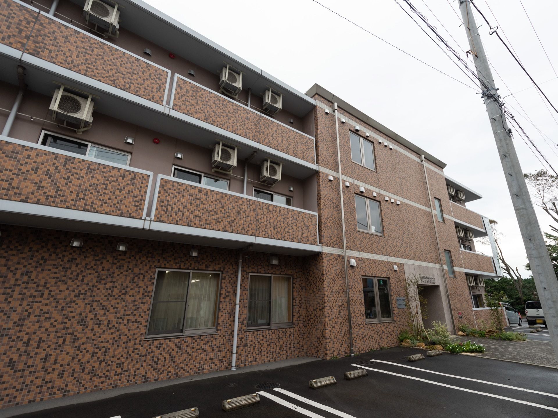色違いのレンガ積み模様の外壁を持つ３階建ての施設である。玄関横の敷地内に白線で仕切りが描かれた駐車スペースがある。