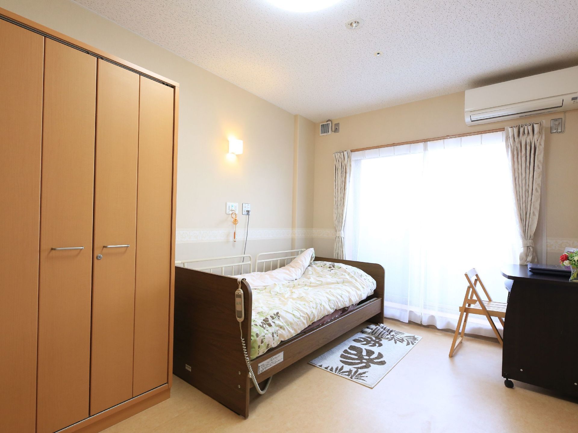 個室タイプの居室で、窓側にキャスター付きのデスクと木製の椅子が置かれている。壁側にベッドが設置されている。