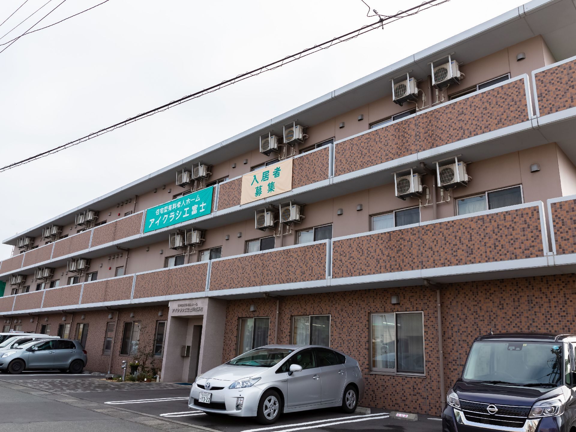 3階建の建物で、ベランダ部分に施設名が書かれた看板が設置されている。施設前に複数の車が停まっている。