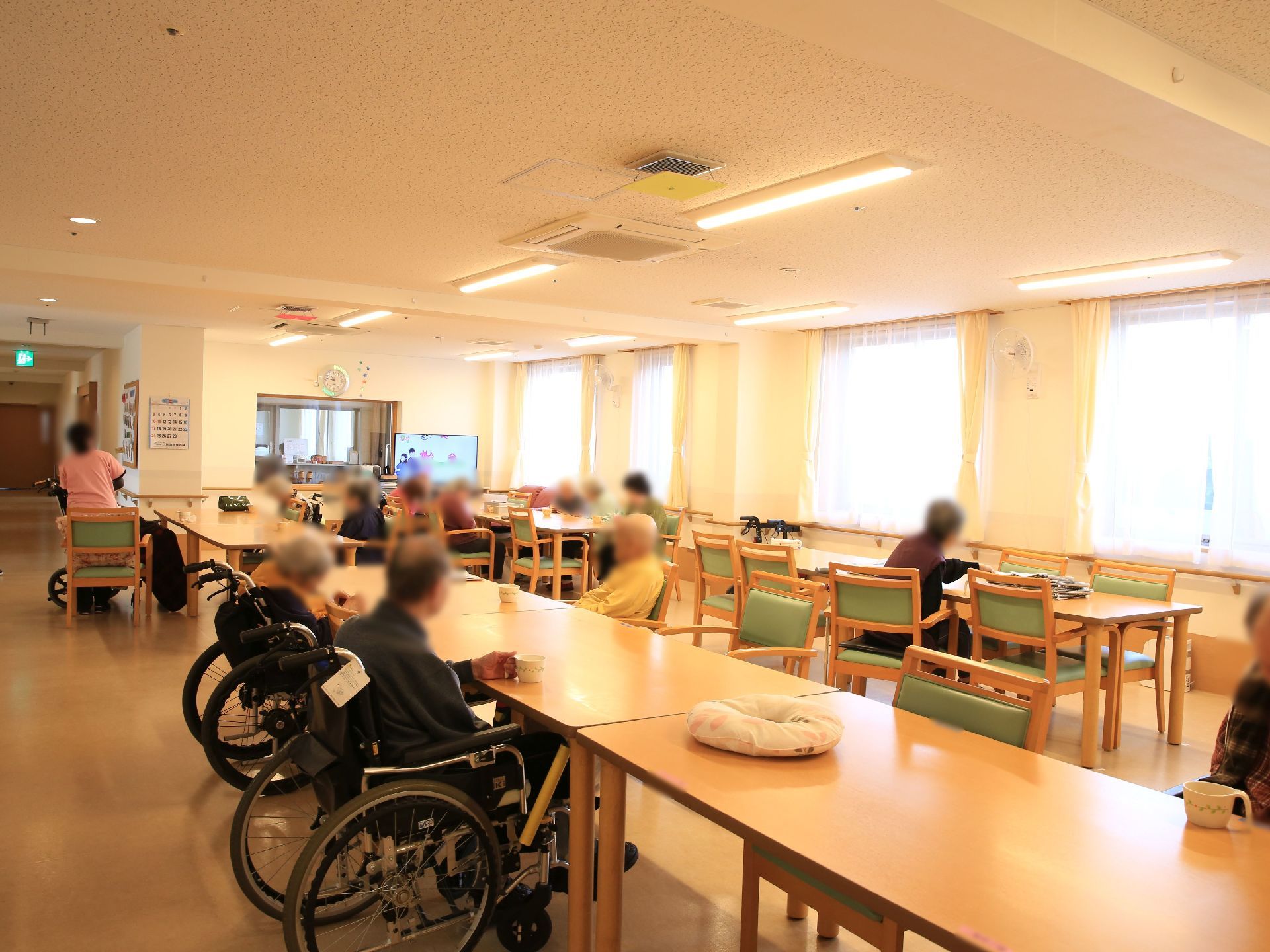 長方形の木製テーブルが並べて設置されており、入居者様が座って過ごされている。スタッフが入居者様がお茶を飲むのを手伝っている。