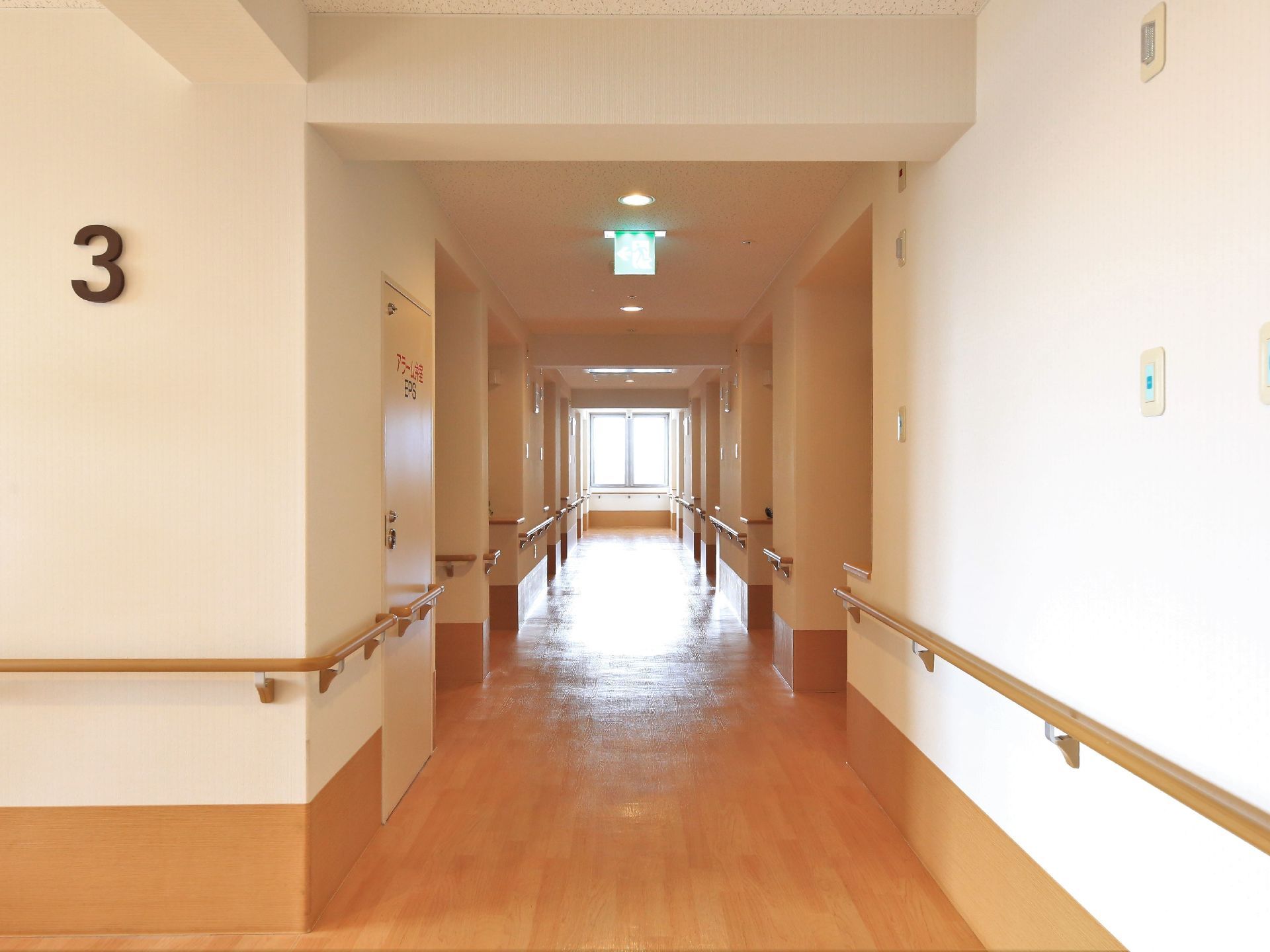 壁に3というマークが書かれた直線的な廊下で、突き当たりに窓がある。天井に避難誘導灯が設置されている。