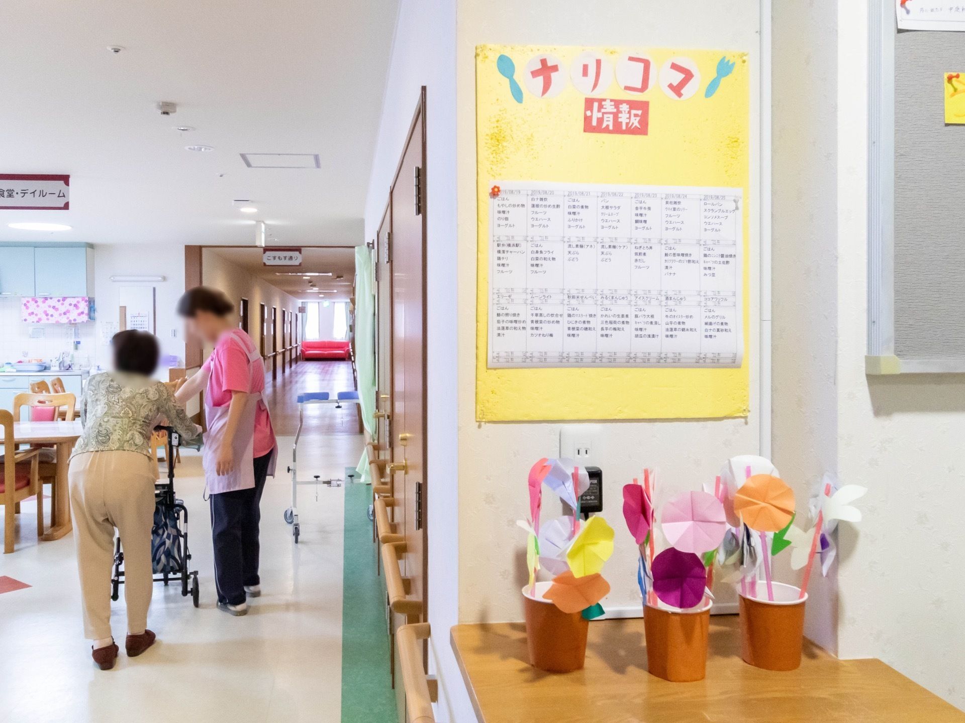 通路の一角に、折り紙で作られた鉢植えのアサガオを模した飾りが置かれている。壁には情報誌が貼られている。
