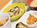 食事のおかずがアップでうつされている。手前には皮のついた焼き魚と大根おろし、奥には肉じゃがとワカメなどのサラダがトレイに乗せられている。