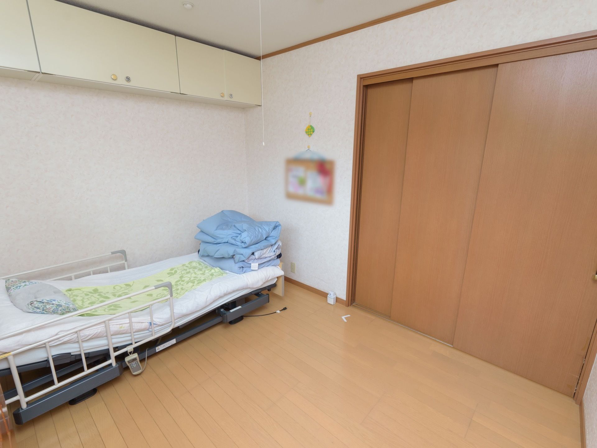 広い洋風の居室。介護用のベッドが置かれていて、ベッドの上の天井付近には、作り付けの戸棚が設置されている。右手には引き戸の扉があるのが見える。