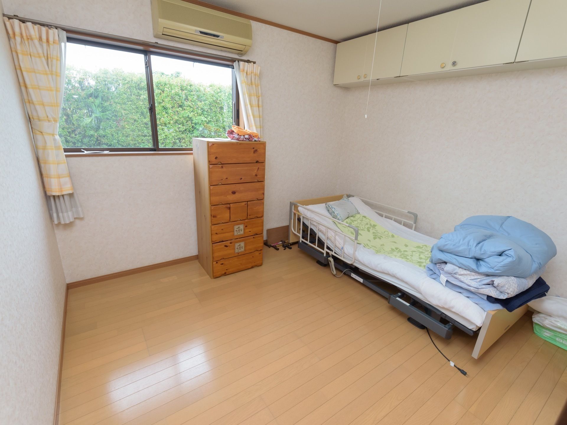 部屋の正面に窓があり、窓の前にはタンスが置かれ、窓上方にはエアコンが設置されている。右手側にはベッドがあり、ベッドの上方には収納棚がある。