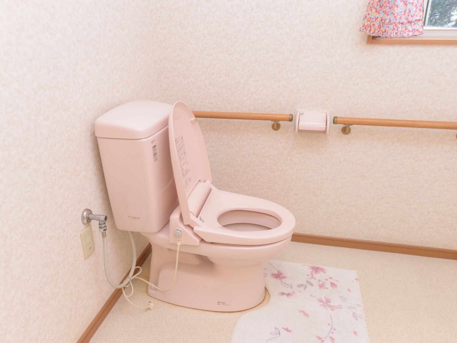 トイレの個室に、淡いピンク色の洋式の便器が設置されている。壁には手すりが設置され、床にはトイレマットが敷かれている。