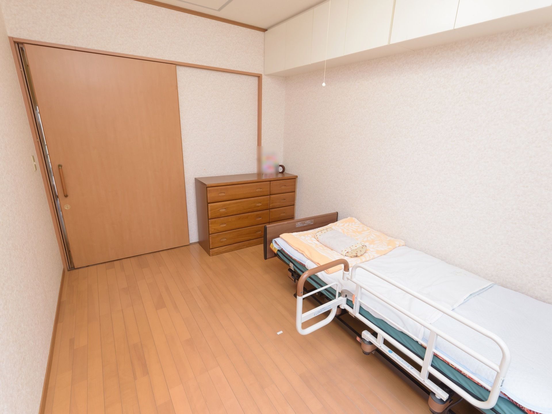 引き戸の大きな扉のある、洋風の居室。室内には介護用のベッドが設置され、収納用のタンスも置かれている。