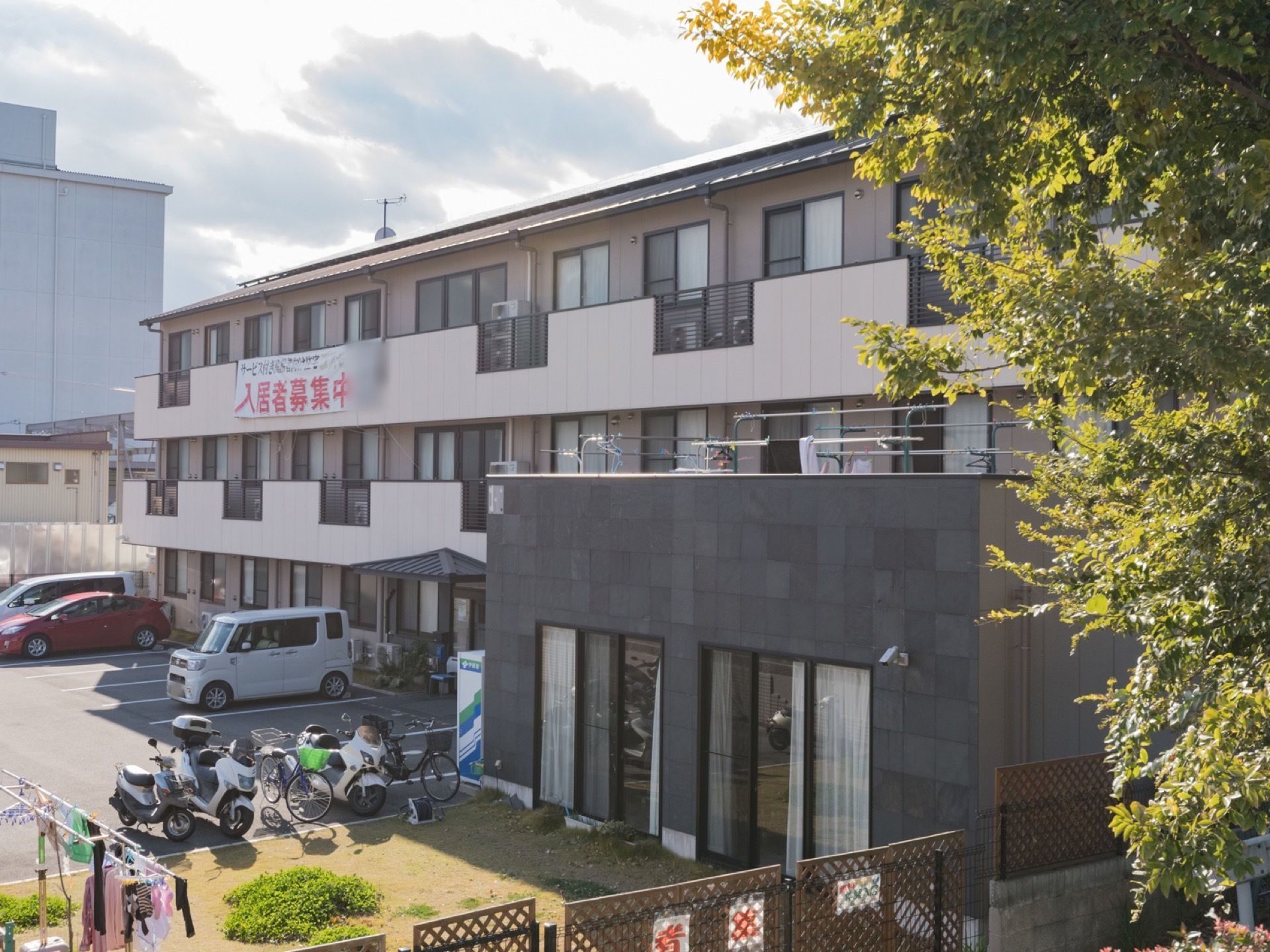 施設は３階建てである。駐車スペースには自動車が複数停まっている。バイクと自転車も置いてある。洗濯物が干してある。