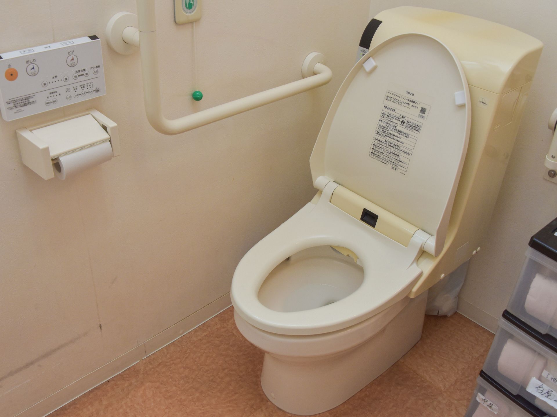 施設のトイレはウォシュレットの水洗トイレ。壁にスイッチとペーパー、手すりが設置されている。便器の横には可動式の手すりと備品が置かれている。