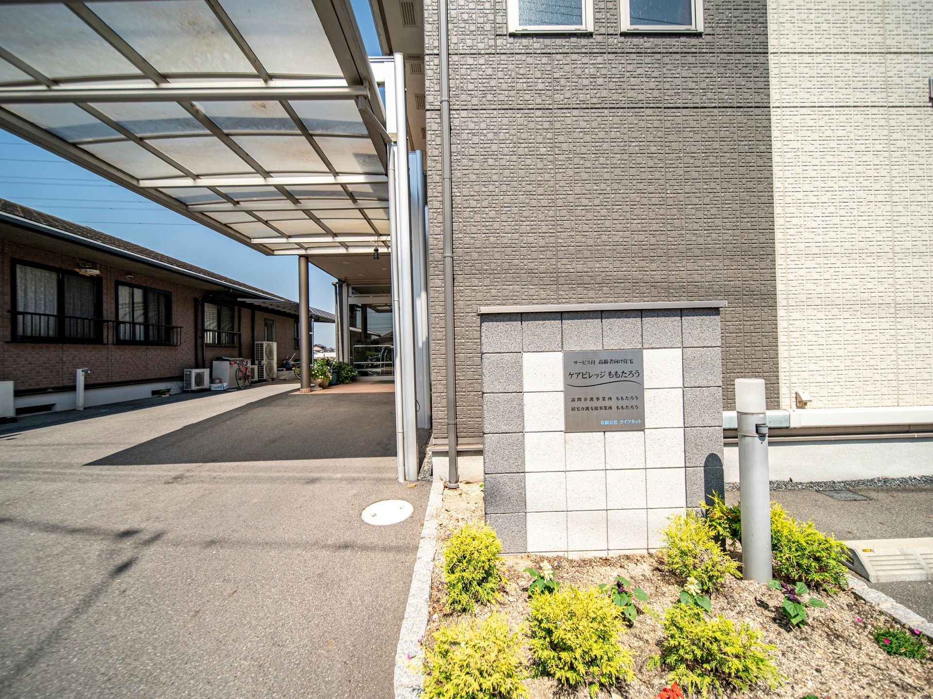 グレー色が基調の大きな建物である。玄関に続く通路に屋根があり、濡れずに移動することができる。建物の前には施設名が表示されている。