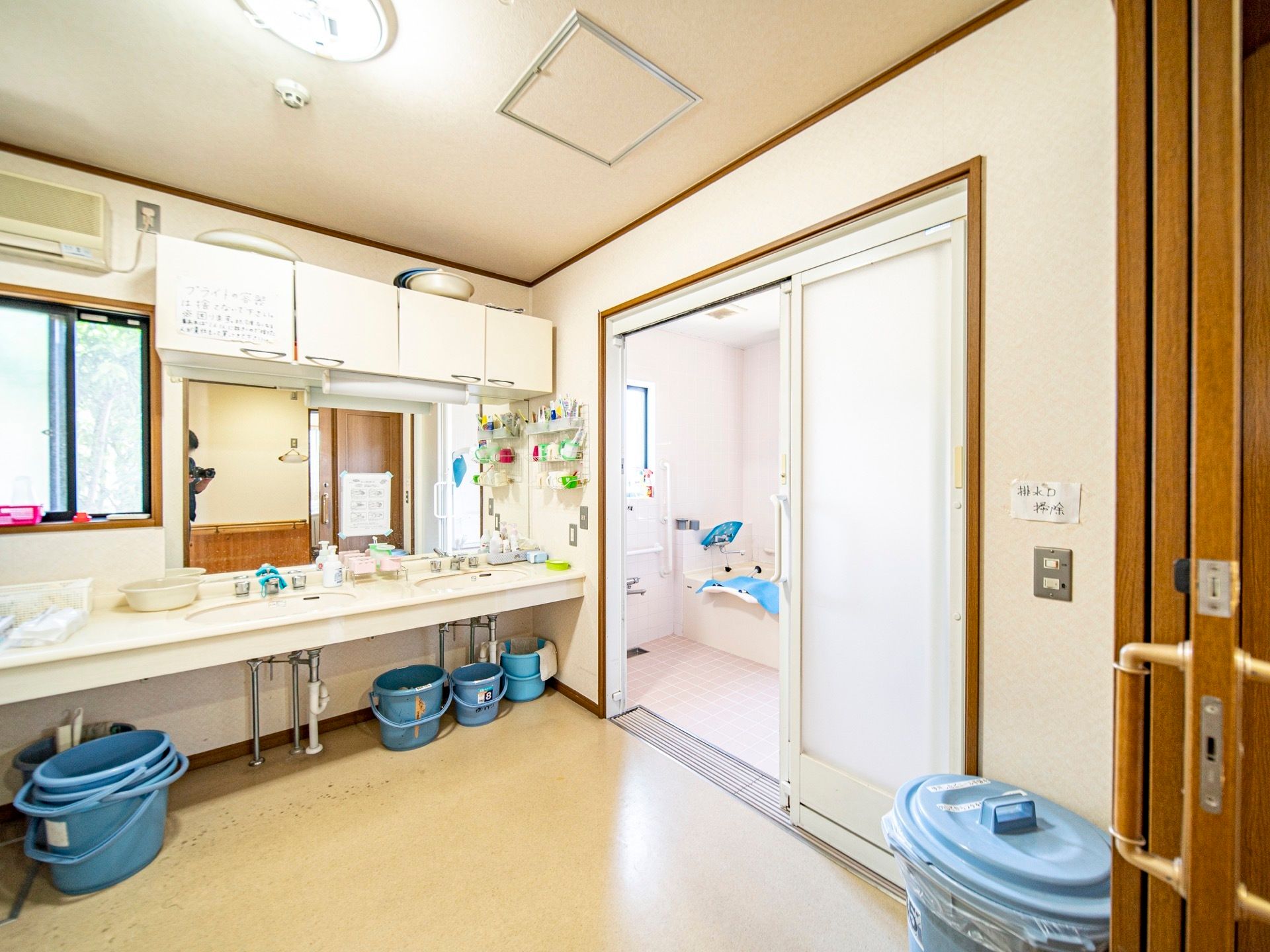 広々とした洗面脱衣所である。エアコンが完備されており、車椅子対応洗面台が設置されている。複数のバケツが置かれている。