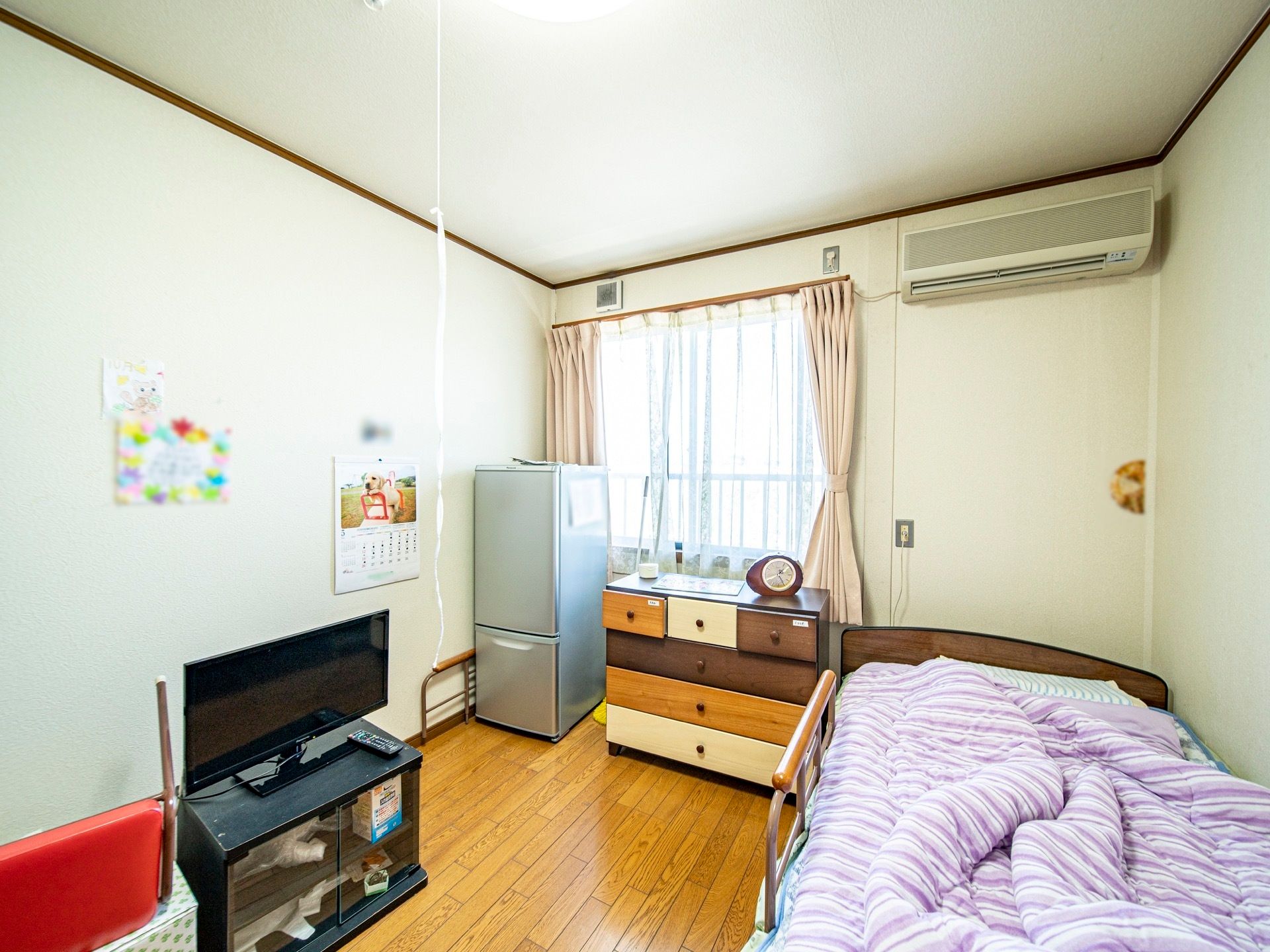 部屋に冷蔵庫やテレビが設置されている。壁にはカレンダーが貼られており、収納棚の上には目覚まし時計が置かれている。