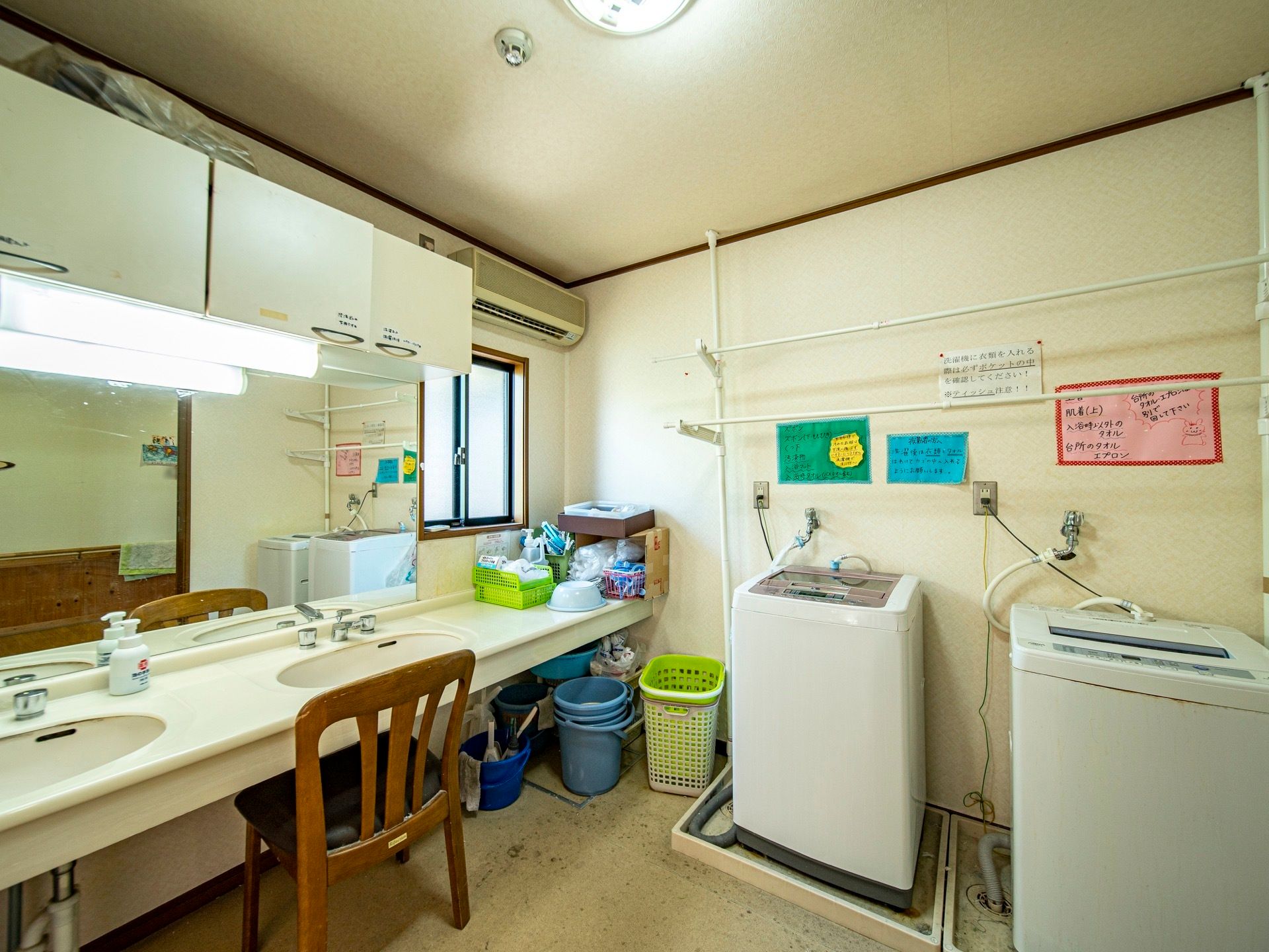 洗面台の前に椅子が並べられている。洗濯機が置かれており、壁には注意事項が書かれた貼り紙が貼り出されている。