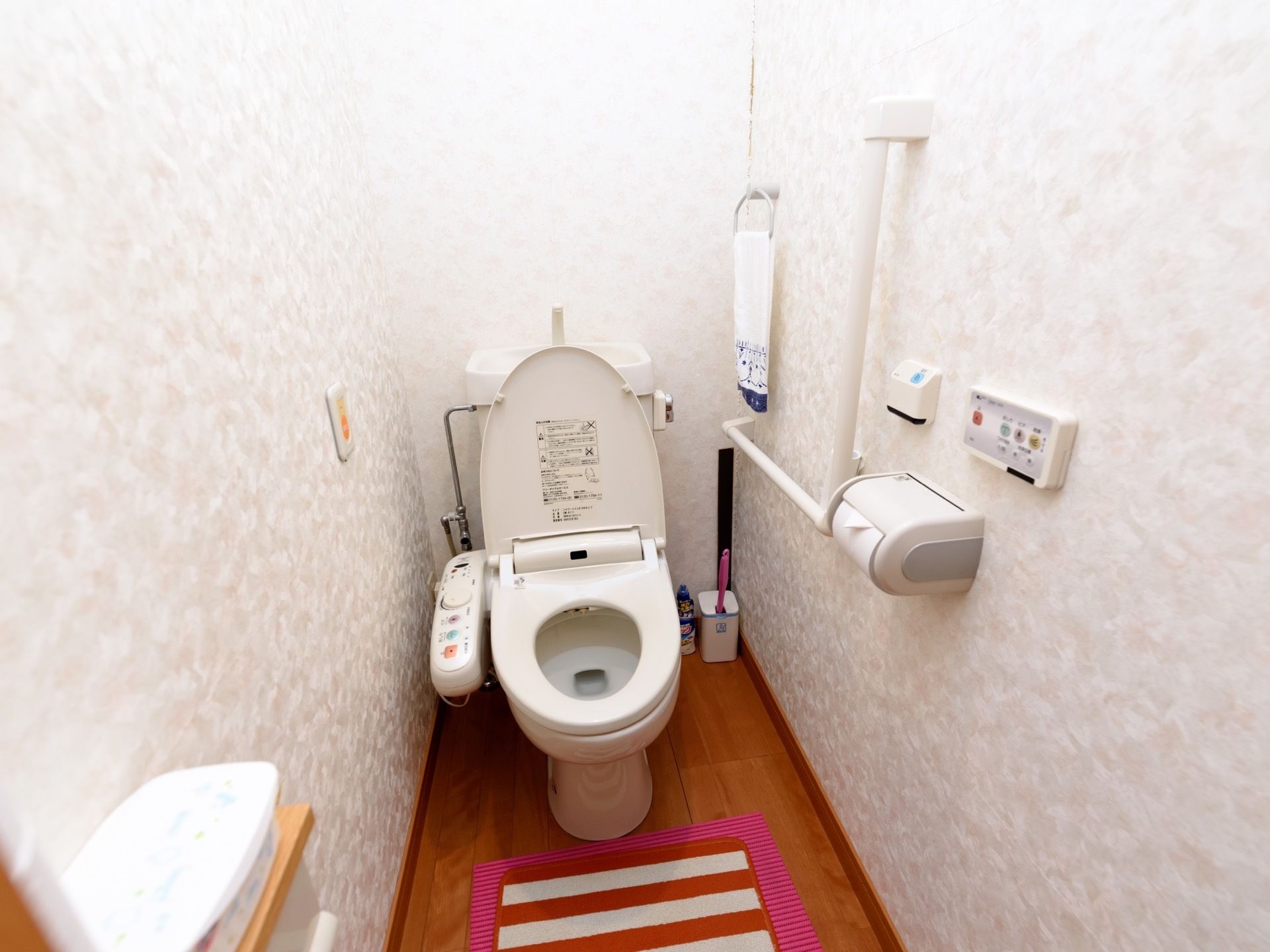 木目調のフローリングに模様入りの壁のトイレスペースである。便座横の壁にエル型の手すりやタオル掛けなどを備えている。