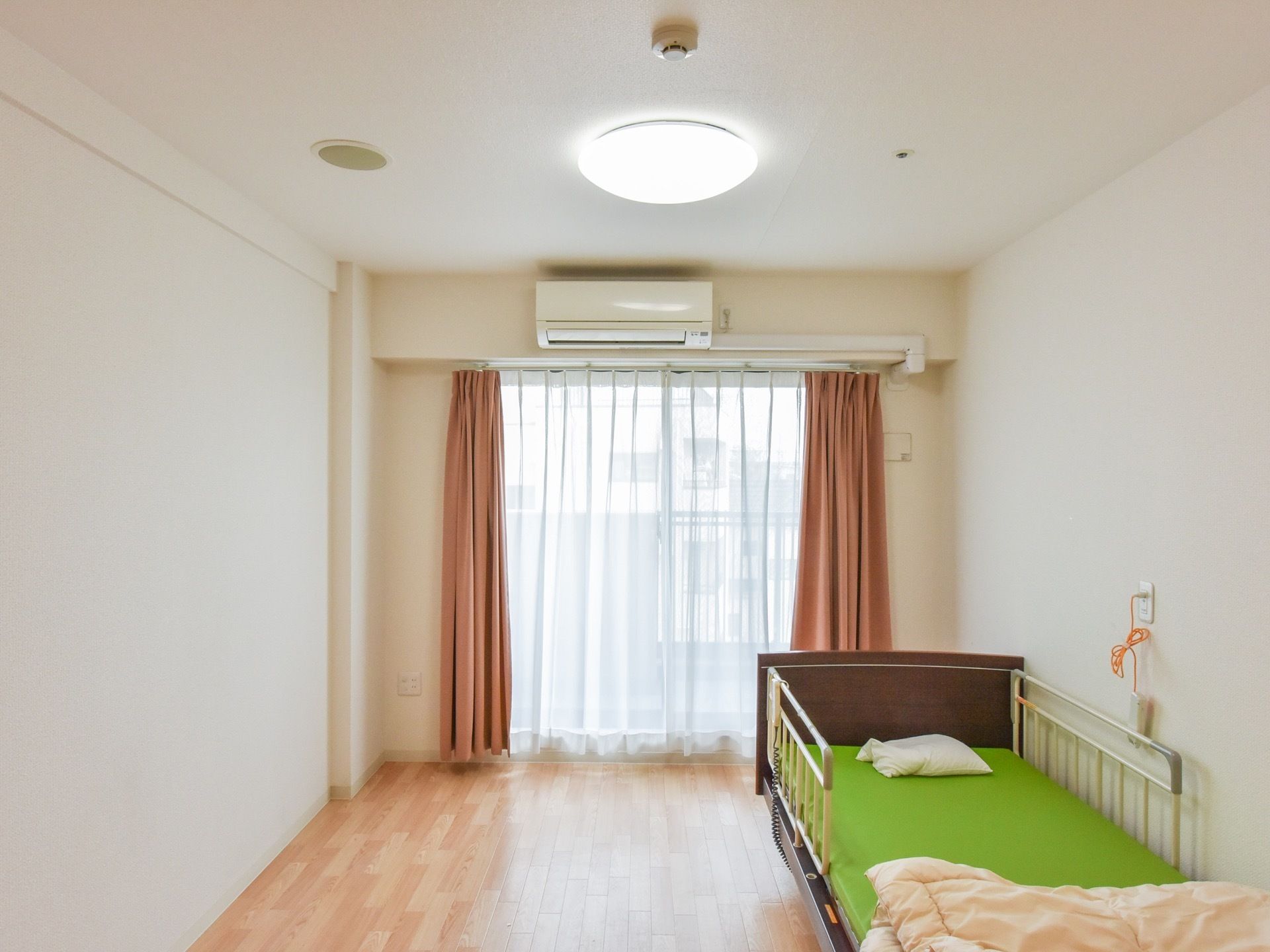 居室には大きな掃き出し窓があり、白いレースとベージュ色のカーテンが掛けてある。その上に、エアコンが設置されている。
