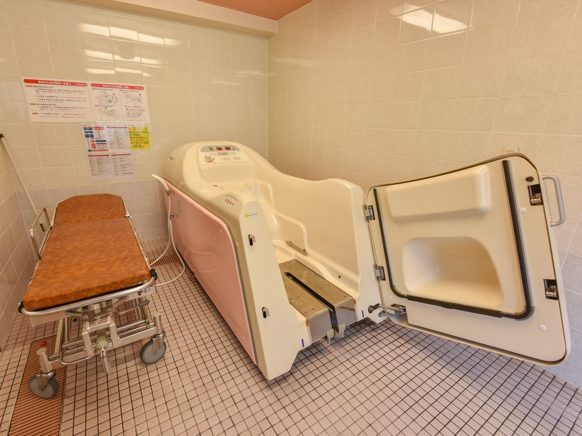 介護入浴機器は、ペールピンクと白が組み合わされた優しい色合いでデザインされている。左横に、ストレッチャーがある。
