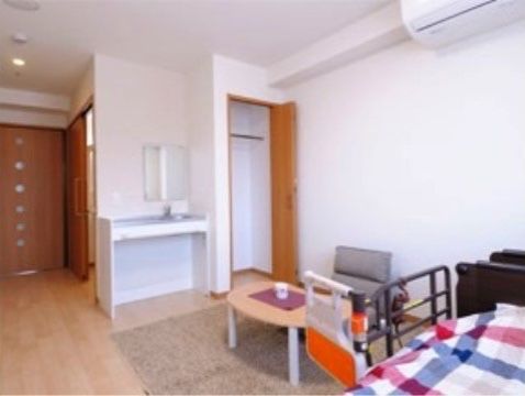 居室奥の窓側から入口を見た様子である。ハンガーバー付きのクローゼット、洗面台、エアコンが取り付けられ、介護ベッドが置かれている。