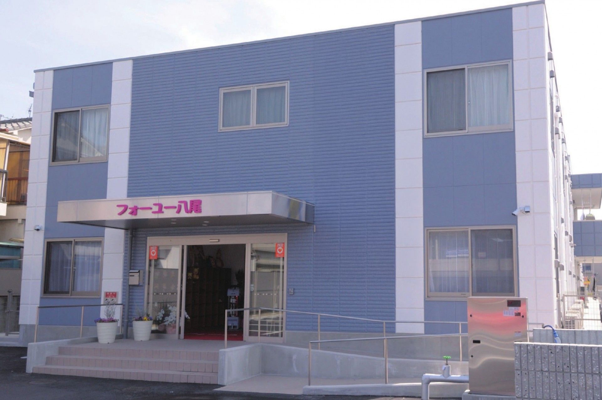 青を基調とした２階建てのグループホームである。入口はガラス張りの自動ドアで、前に手すりが設置されたスロープや階段がある。