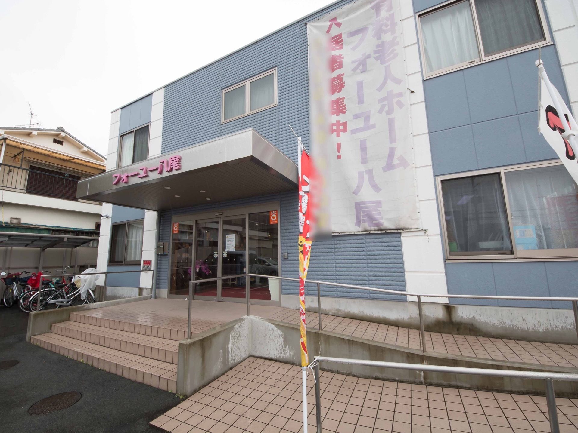 青を基調とした２階建てのグループホームである。入口はガラス張りの自動ドアで、前に手すりが設置されたスロープや階段がある。
