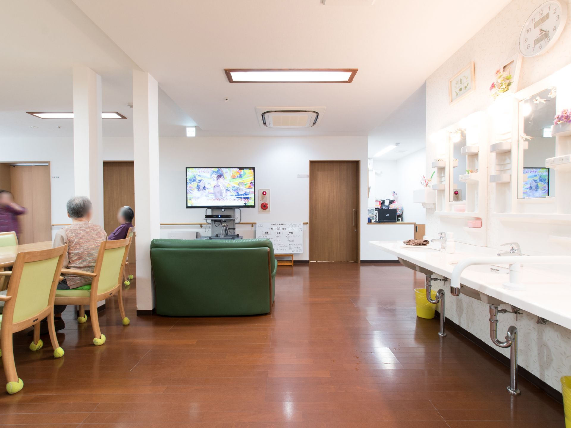テレビの前には緑色のソファーもあり、座ってテレビをみることもできる。洗面所が近くにあり、棚には花などが飾られている。