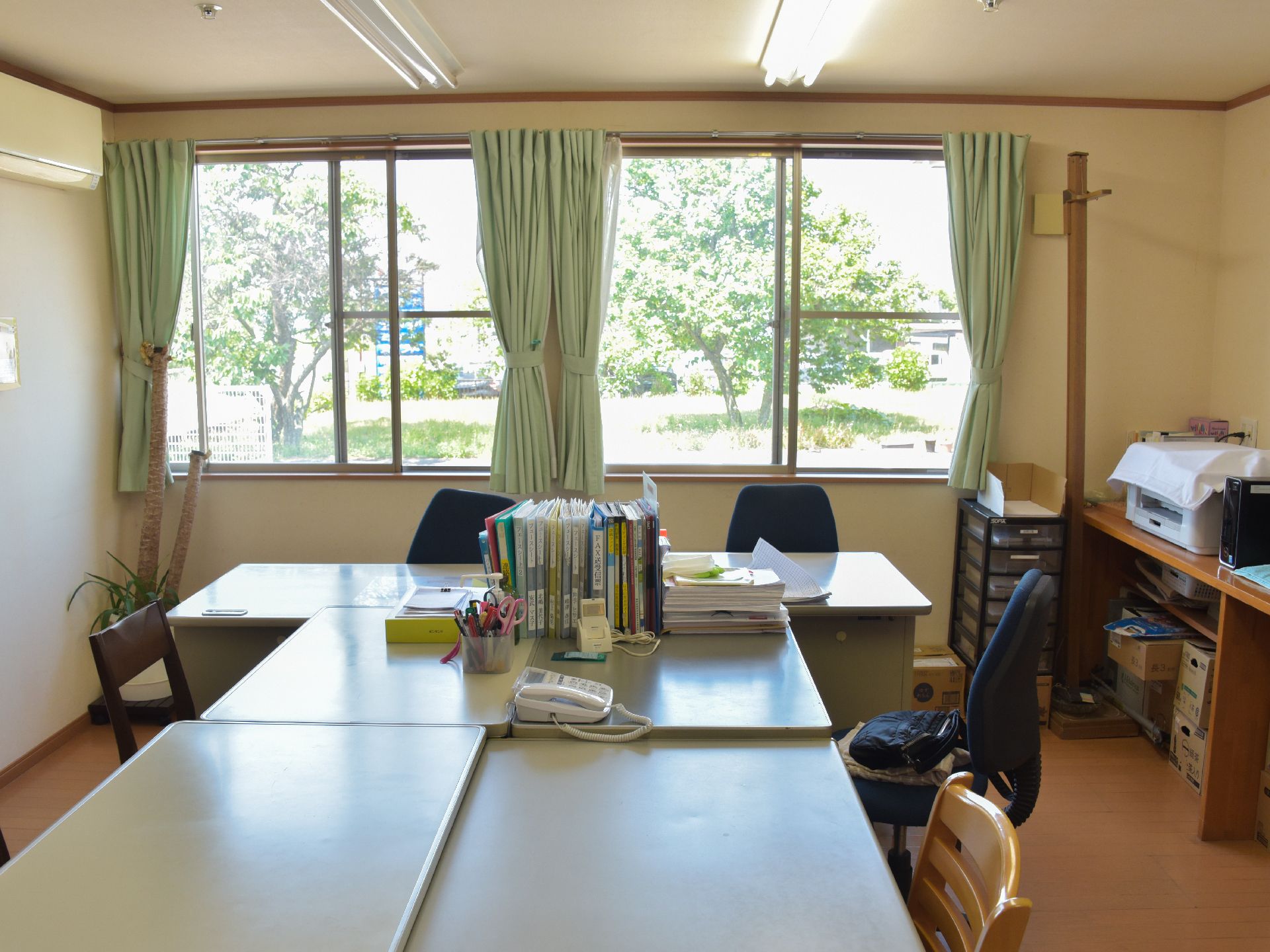 事務所は庭に面していて窓からは外の景色がよく見える。中央に置かれたデスクには電話やファイル、備品などが置かれているのが見える。