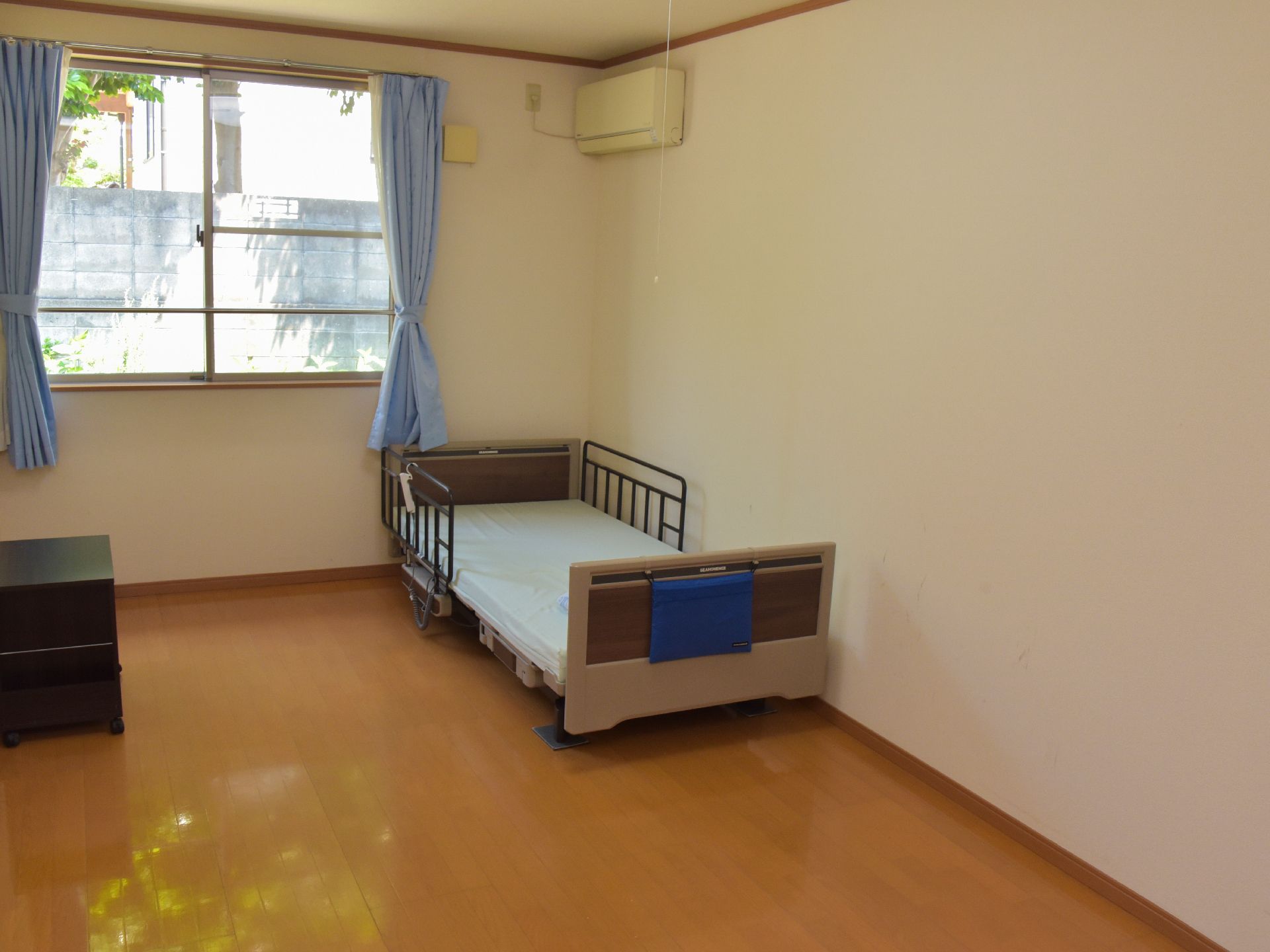 居室は白壁に茶色のフローリング床。奥には窓がありカーテンもある。壁際に介護用のベッドが一台設置されている。エアコンも備え付けられている。