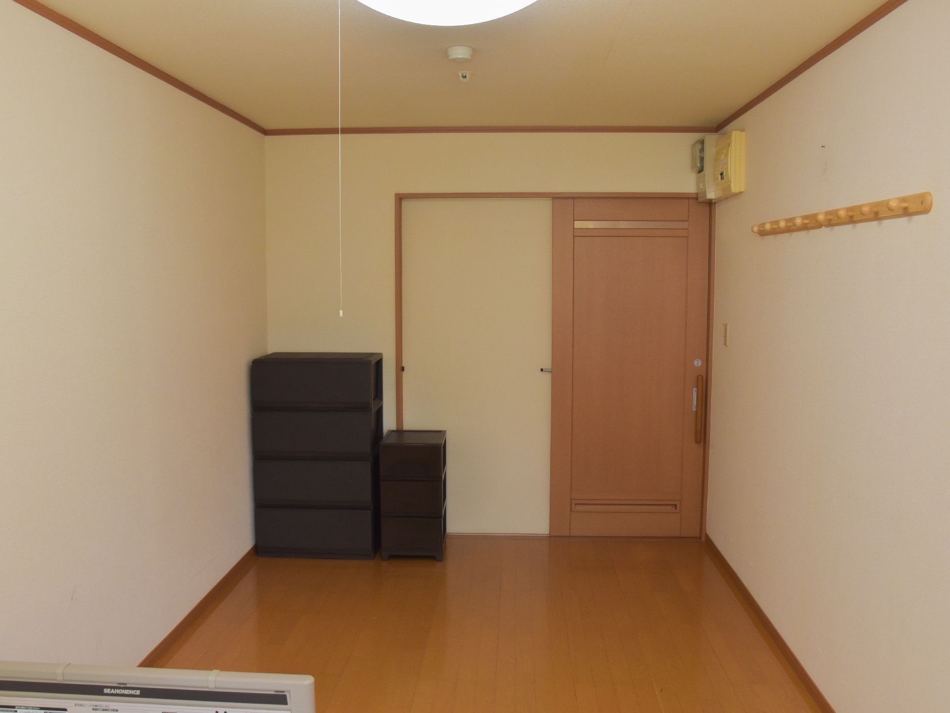 居室の扉は茶色の木目調で横にスライドするタイプになっている。入口近くにはブレーカーがあり、服を吊るすフックもある。黒いケースも置かれている。