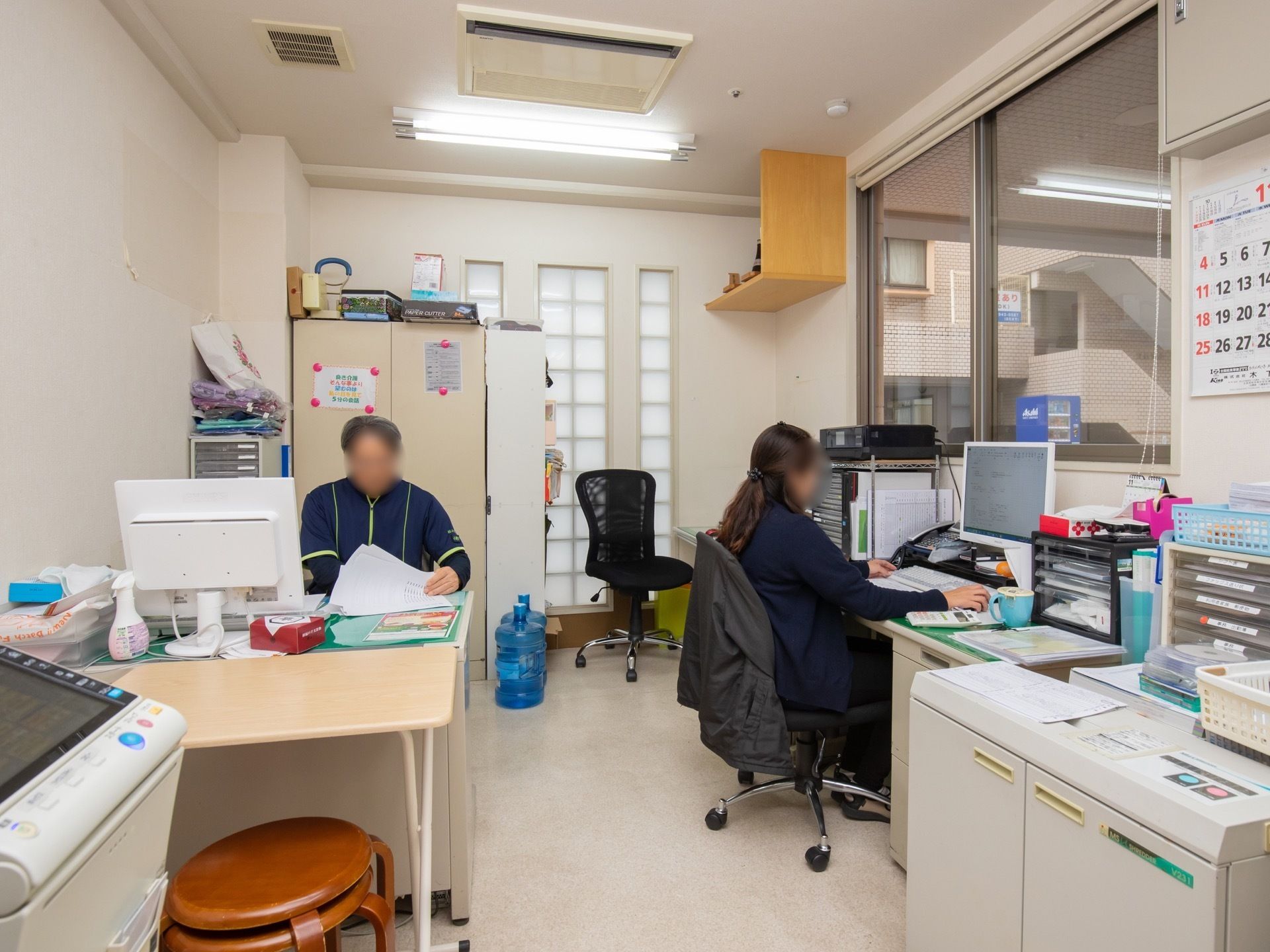 事務所には、入居者様が外から声を掛けやすいよう、大きなガラス窓が備わっている。２名のスタッフがパソコンの前で事務を行っている。