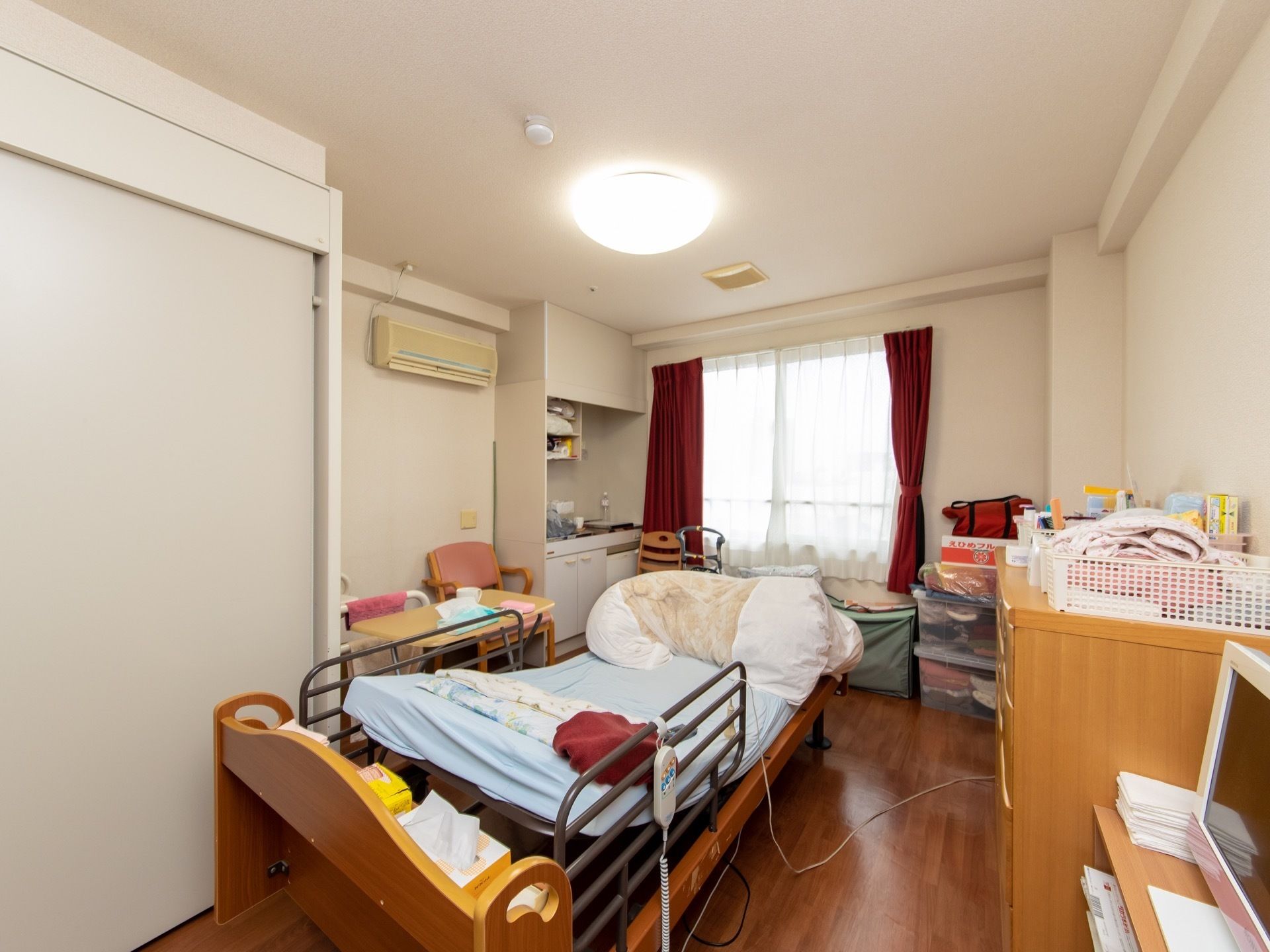 白壁と床がフローリング仕様の明るい居室である。介護用ベッドに簡易キッチン、机と椅子、収納棚が備わっている。