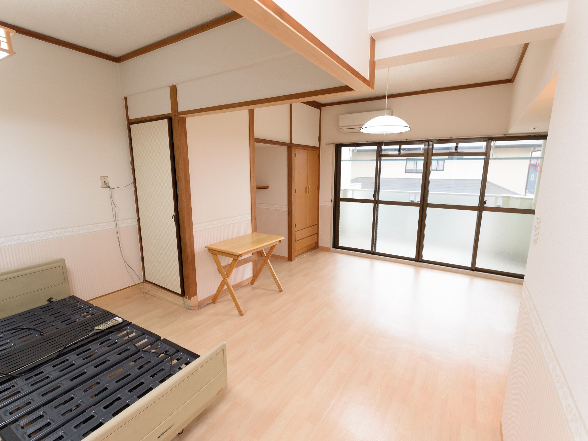 太陽光の入る大きな窓がある。部屋にはベッド、エアコン、クローゼット、テーブルなどの家具が設置されている。