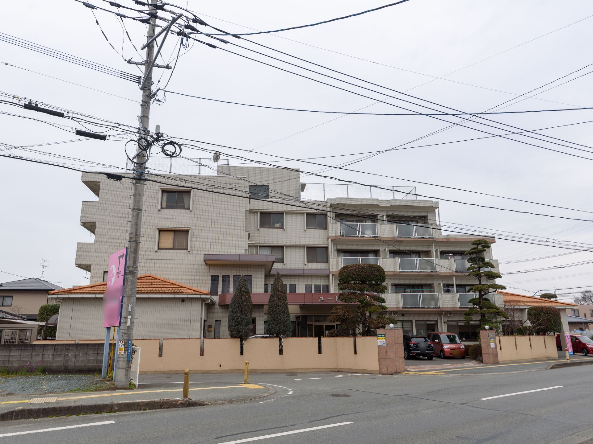 道路に面した場所に建つ５階建てのベージュの建物である。高い塀があり、敷地内には駐車スペースが設けられている。