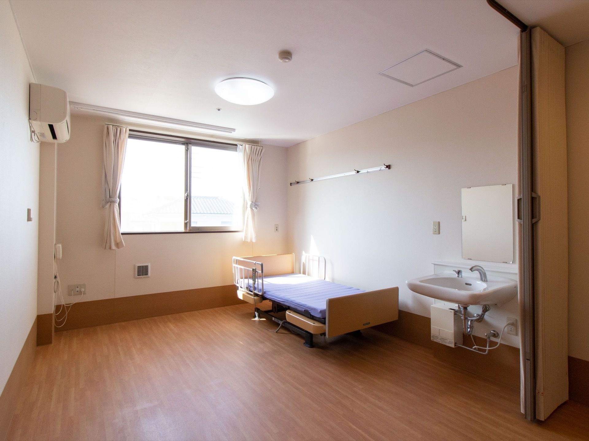 腰高の窓がある居室である。壁際に介護用ベッドが置かれている。壁には物を掛けられるフックが取り付けられている。