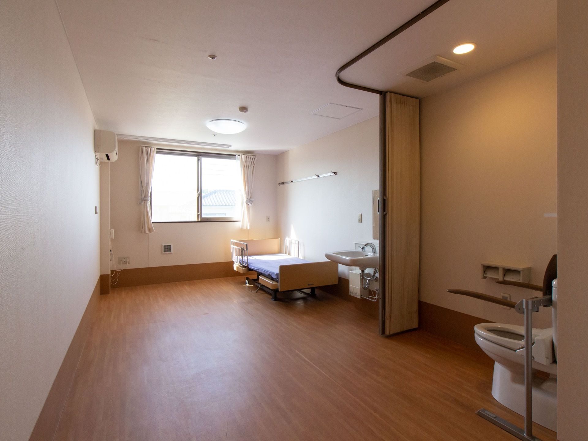 居室の窓際にエアコンが取り付けられている。照明器具は、居間とトイレの天井の２ヶ所に設置されている。介護用ベッドの両側に柵がある。