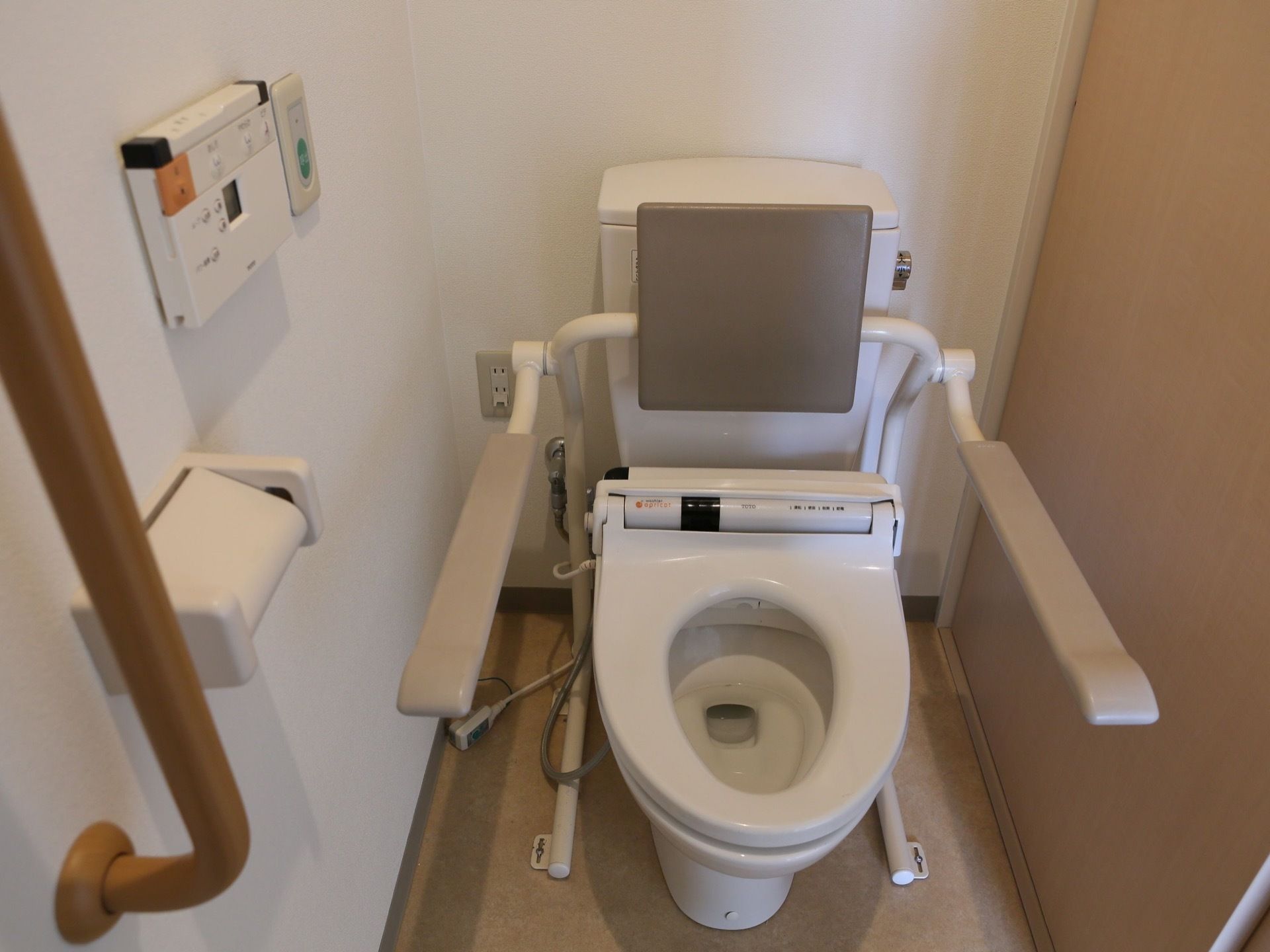 居室内のトイレ。背もたれ、肘掛け式の手すり、縦型手すり、温水便座、緊急呼出しボタンが設置されている。