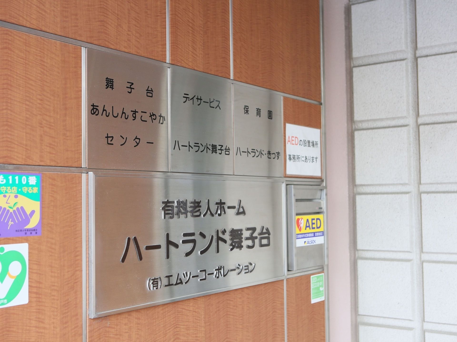 玄関には施設名の看板が掲げられている。デイサービスや保育園などが、施設内に併設されている。AED設置や子供110番などの表示がある。