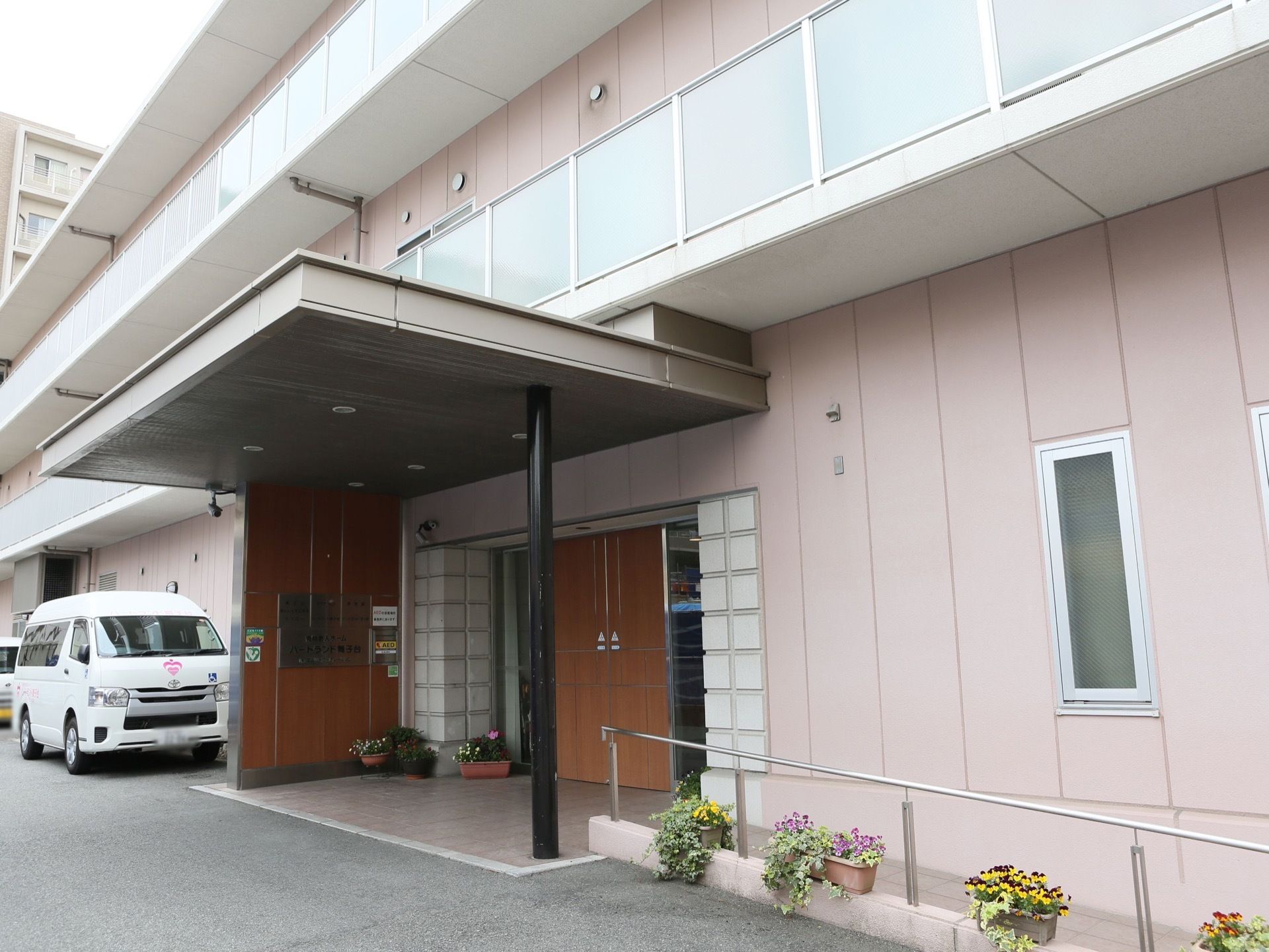 玄関には大きなひさしがかかっている。バリアフリーで段差がなく、スロープと手すりがある。送迎車が玄関前まで進入できる。
