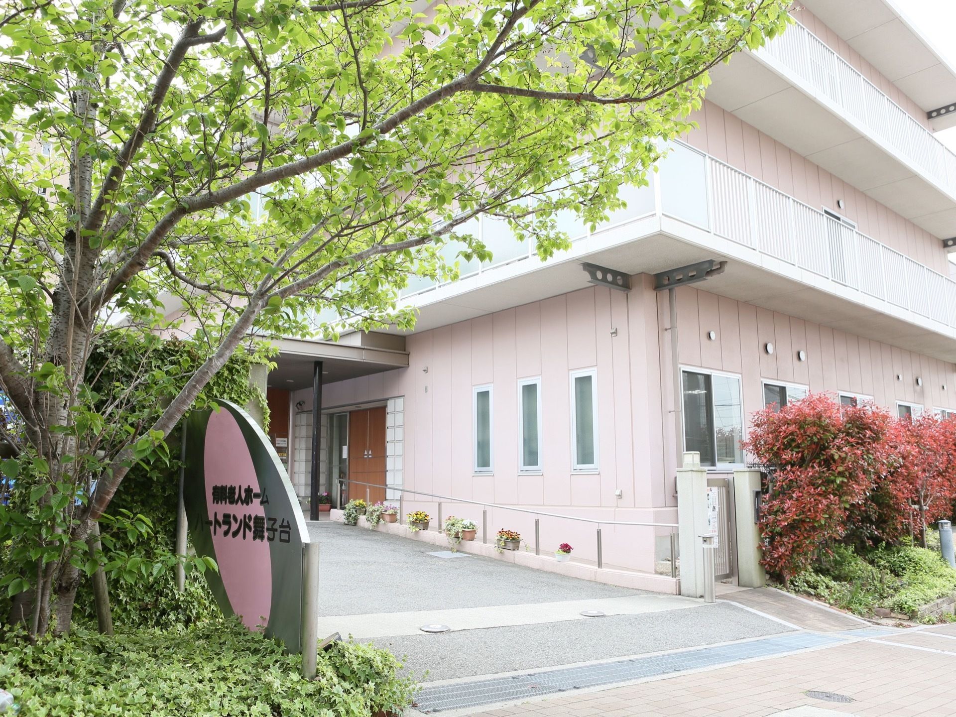 薄いピンクの建物。正面にはさまざまな植物が植えられている。玄関までは緩やかなスロープになっており、手すりが設置されている。