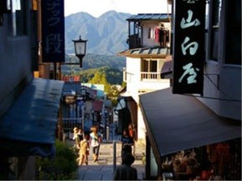温泉街の一角の風景である。坂道の両側に土産物などの店舗がならび、観光客が行き来している。背景には緑の山々が見える。